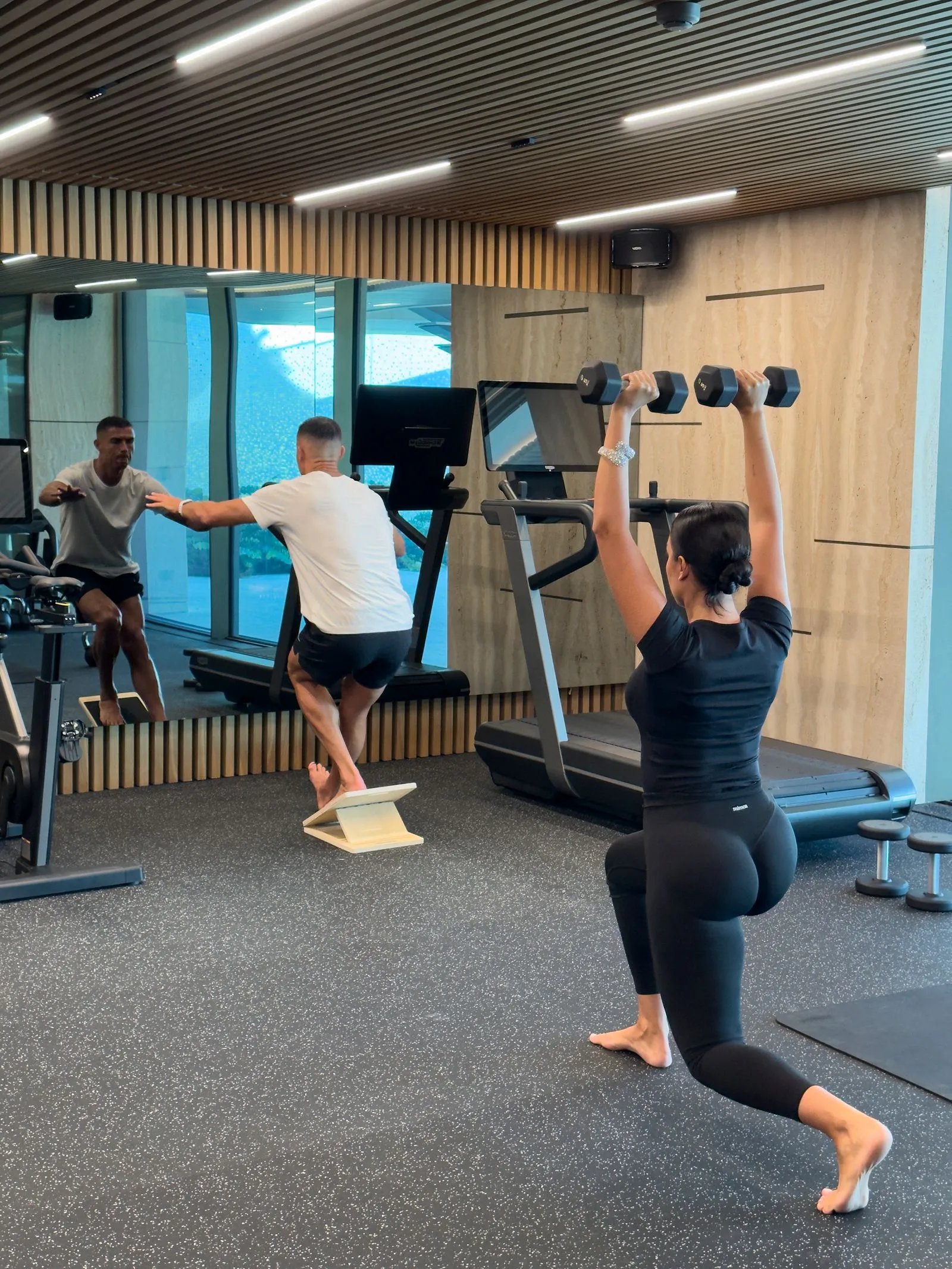 Woman in black athletic wear lunges in a lunge/overhead dumbbell press on a treadmill seen in a gym mirror. Still gym scene with two men exercising nearby.