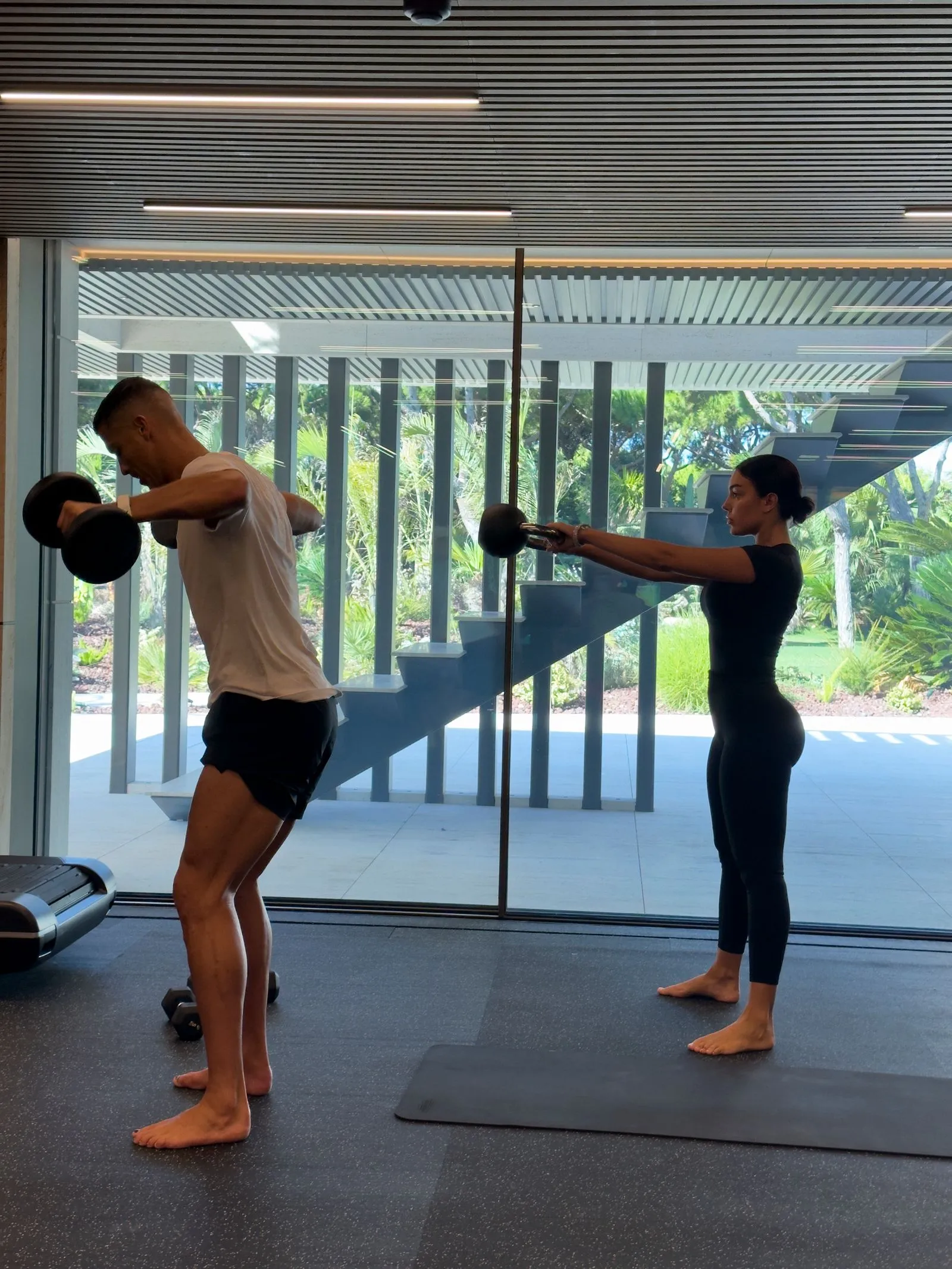 Man bent over with dumbbells performing a row exercise in a gym, woman in black standing behind him holding dumbbells to his side; glass wall and greenery outside.