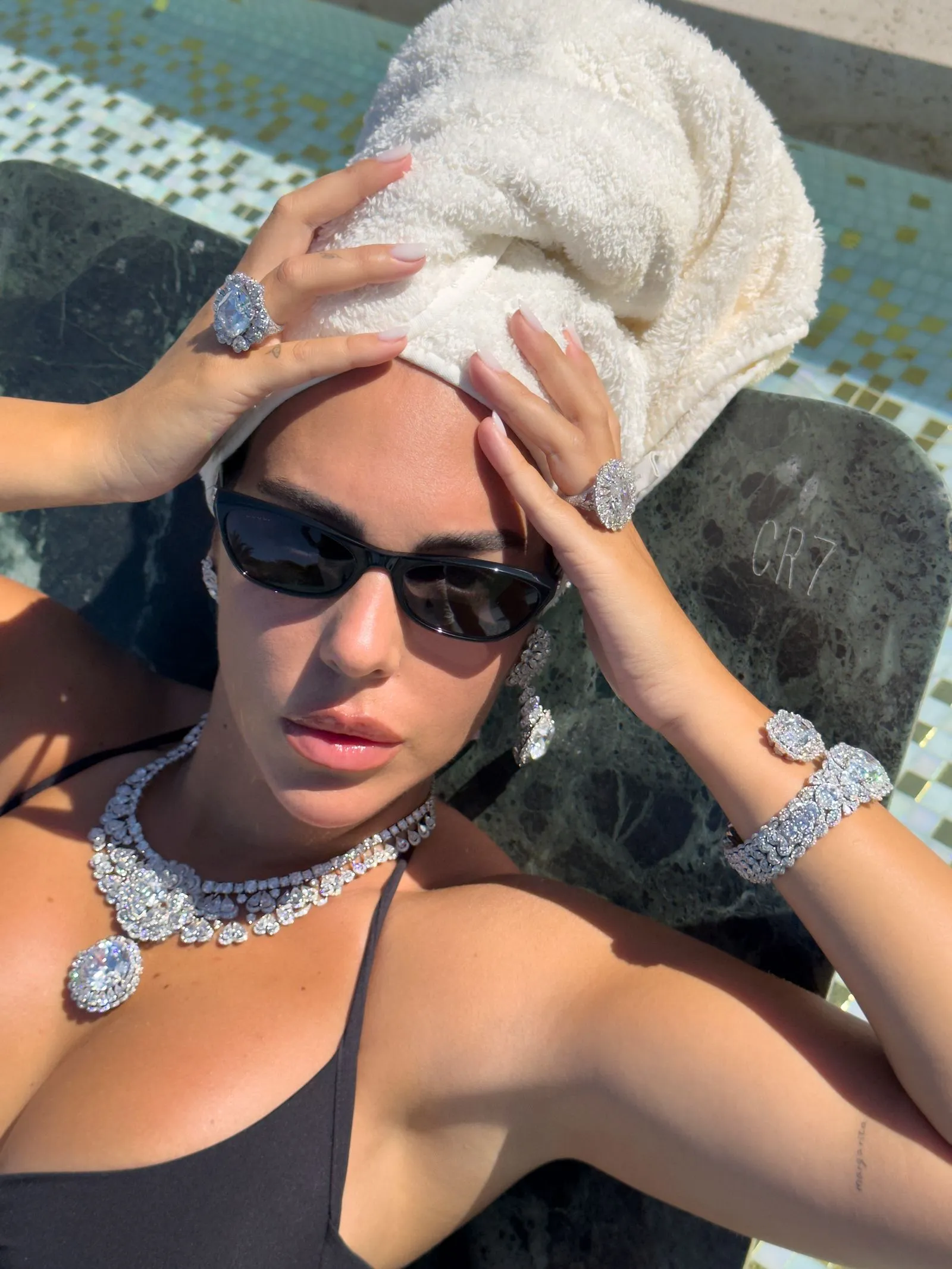 Woman sunbathing by a pool in a black swimsuit, wearing large diamond jewelry and sunglasses, with a white towel wrapped on her head.