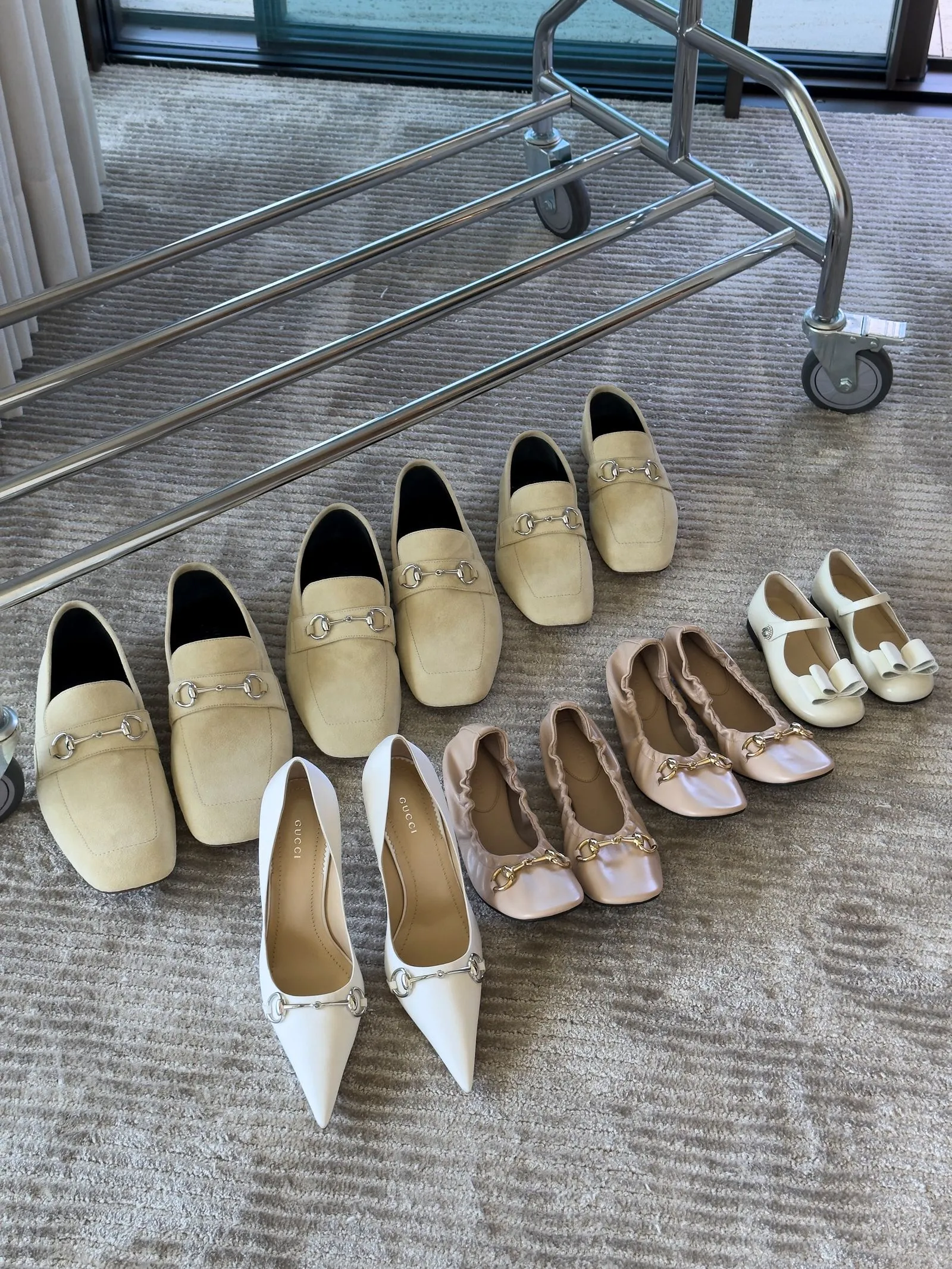 Assorted beige and white women's dress shoes arranged in a semicircle on a textured carpet beside a metal luggage cart with wheels. One white pointed-toe heel shows the Gucci logo on the insole on the left.