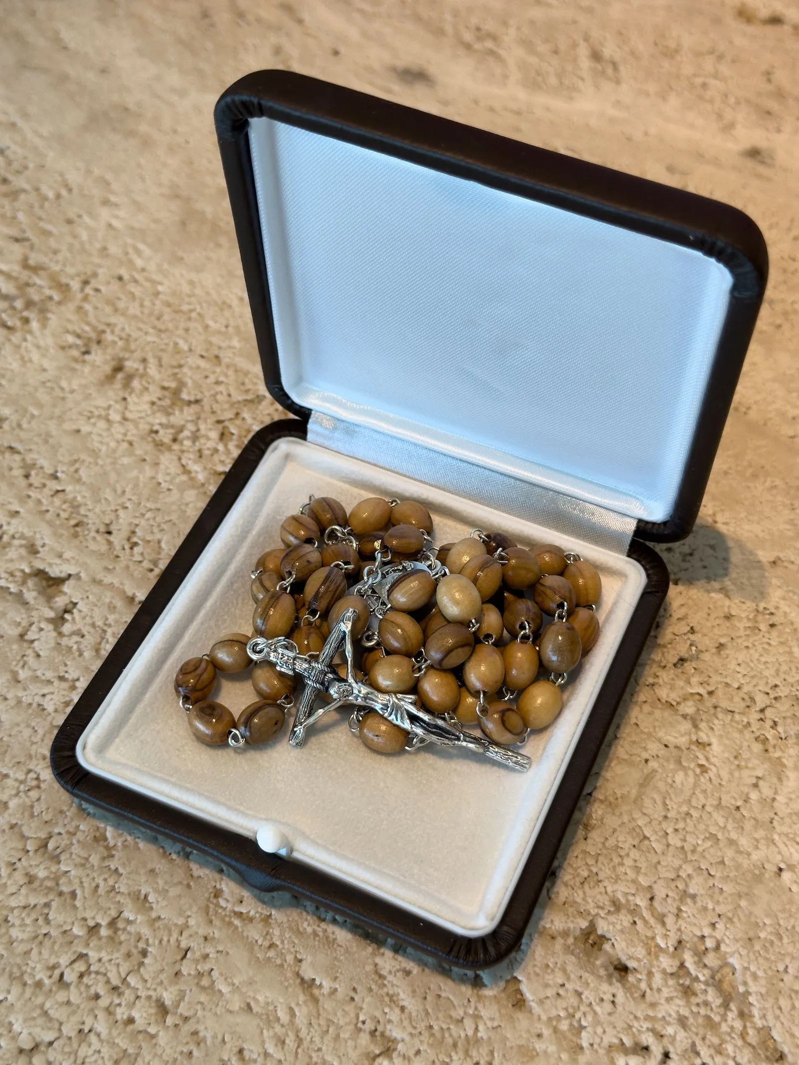 Open jewelry box with wooden bead rosary and metal crucifix resting on a white lining inside the case on a beige countertop.