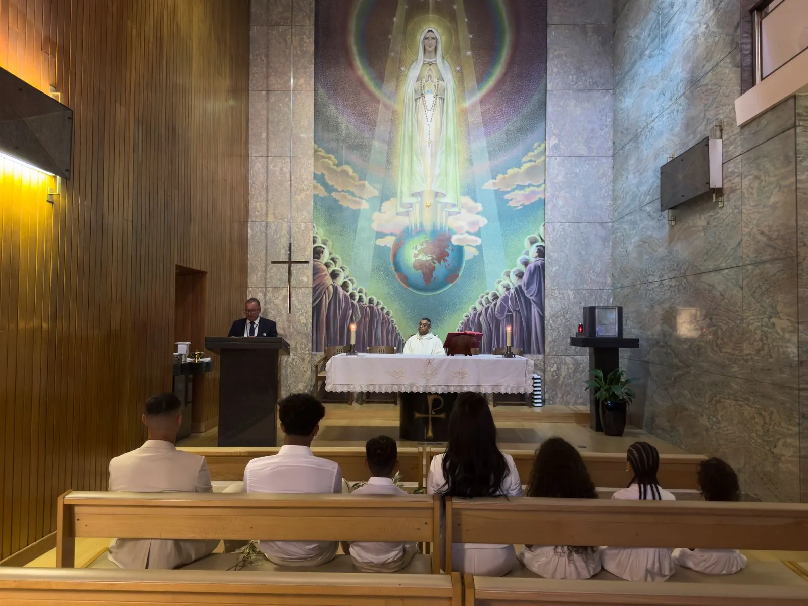 Priest at an altar inside a church, with a large mural of Mary above and worshippers seated.