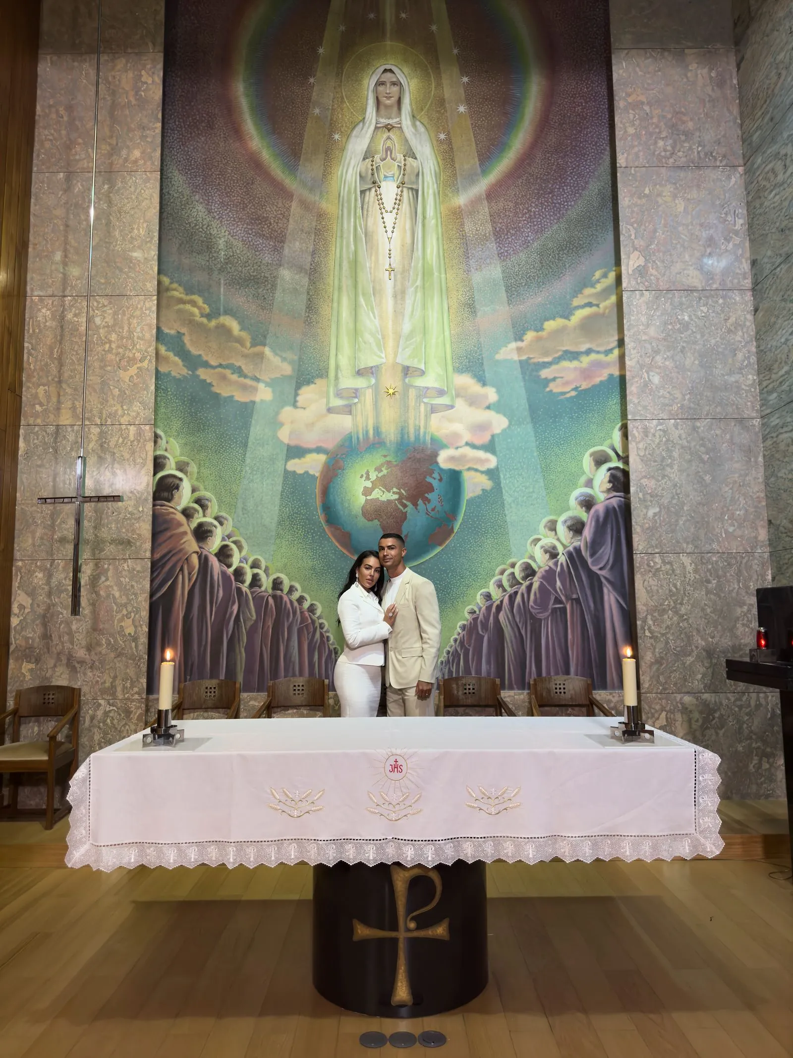 Couple in light-colored attire posing in front of a church altar, with a large mural of the Virgin Mary above and a globe beneath her feet.