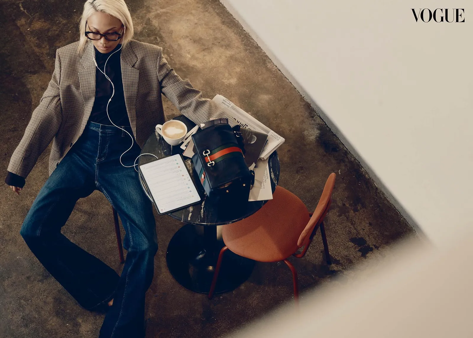 Blonde person in a checkered blazer sits at a round table with a tablet, coffee, backpack, and papers; Vogue logo in the top-right corner.