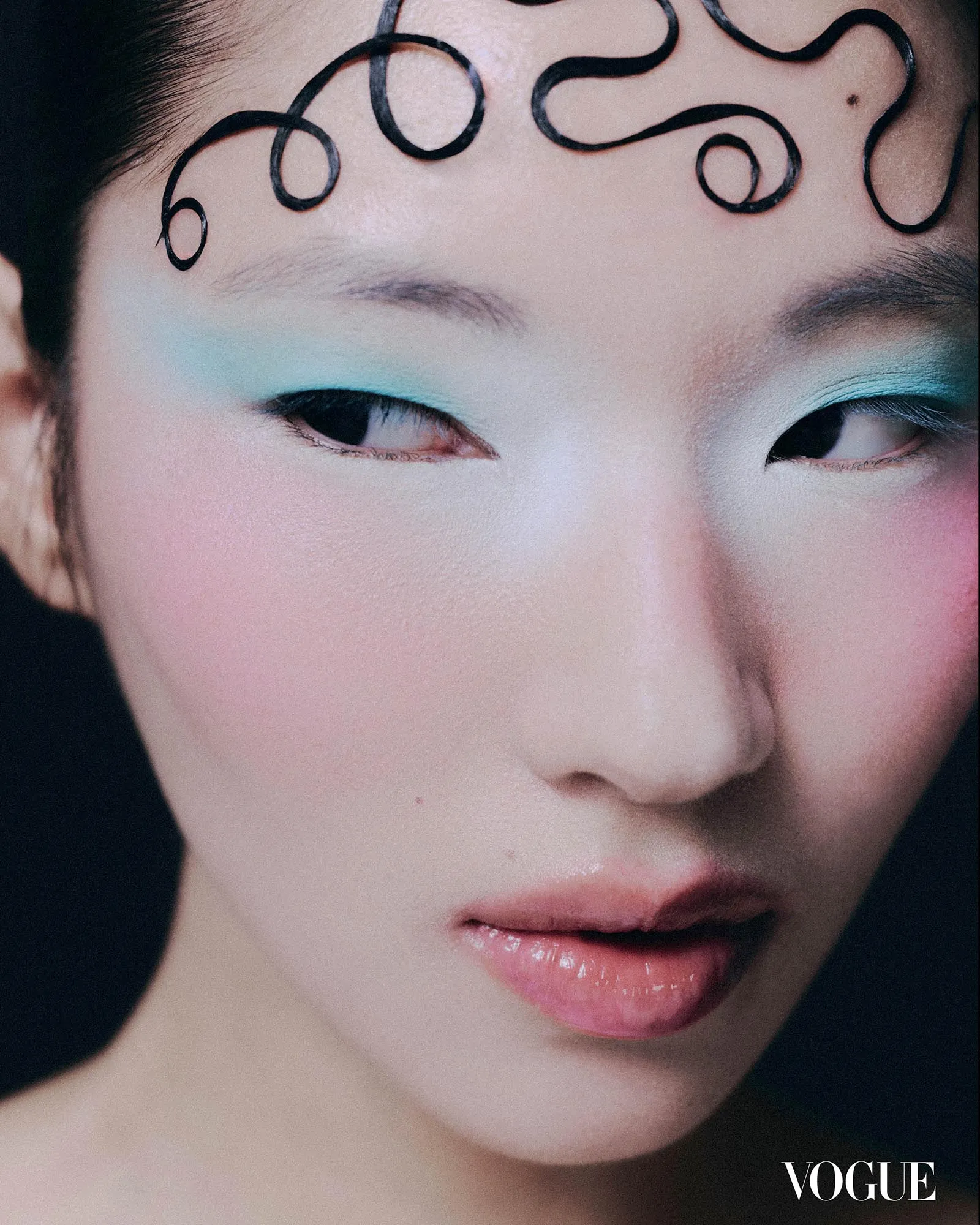 Close-up of a model's face with turquoise eyeshadow, pink lips, and a decorative black curl on the forehead; Vogue logo.