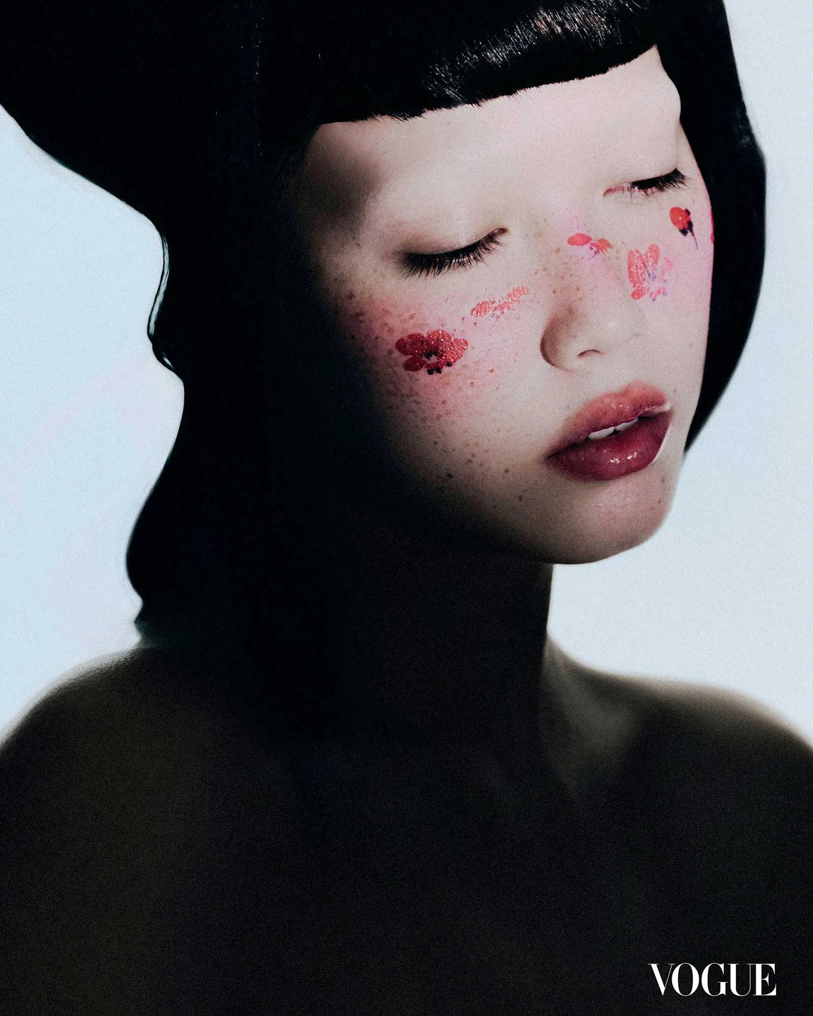 Close-up fashion portrait of a model with a black head wrap, pale skin, pink lips, and red floral face art on the cheek, Vogue logo in the corner.