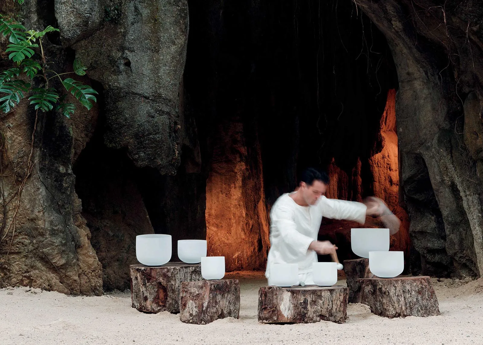 Musician in a white outfit plays white singing bowls set on wooden stumps inside a rocky beach cave.