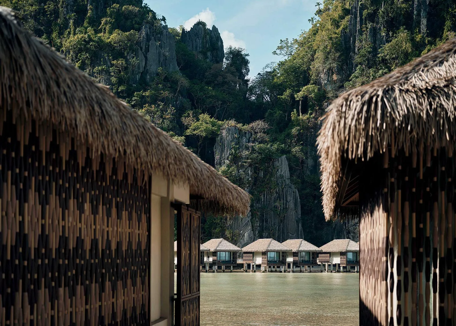 Overwater bungalows with thatched roofs frame a calm lagoon and rocky tropical cliffs in the background.