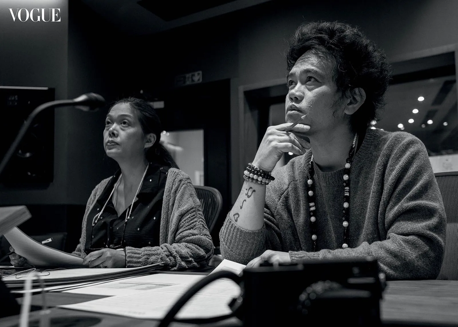 Two women sit at a conference table in a Vogue studio, reviewing papers; the woman on the right rests her chin with a pencil and wears beaded necklaces.