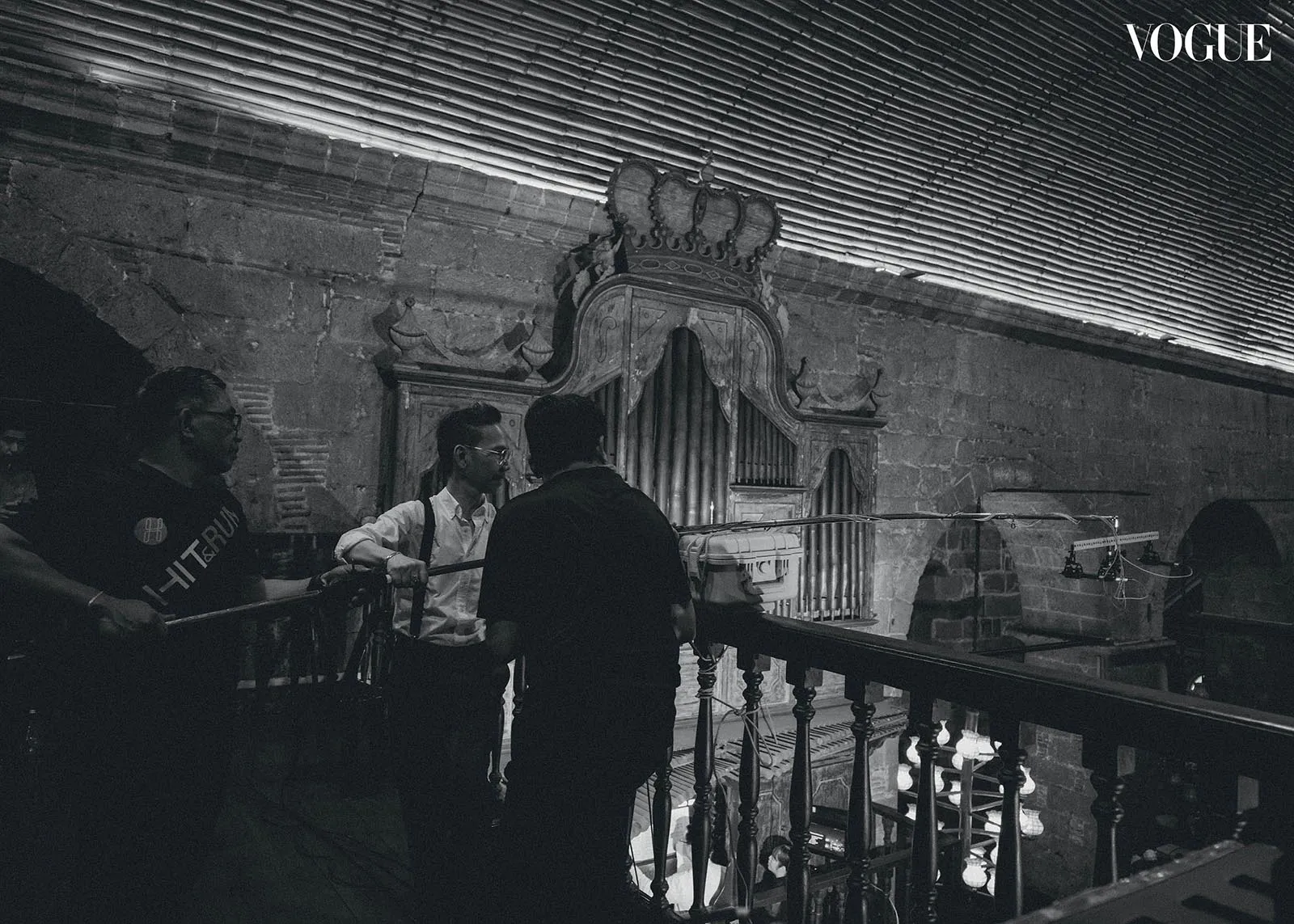 A group of people backstage in a dim stone hall, adjusting a long boom near an ornate organ façade behind a railing.