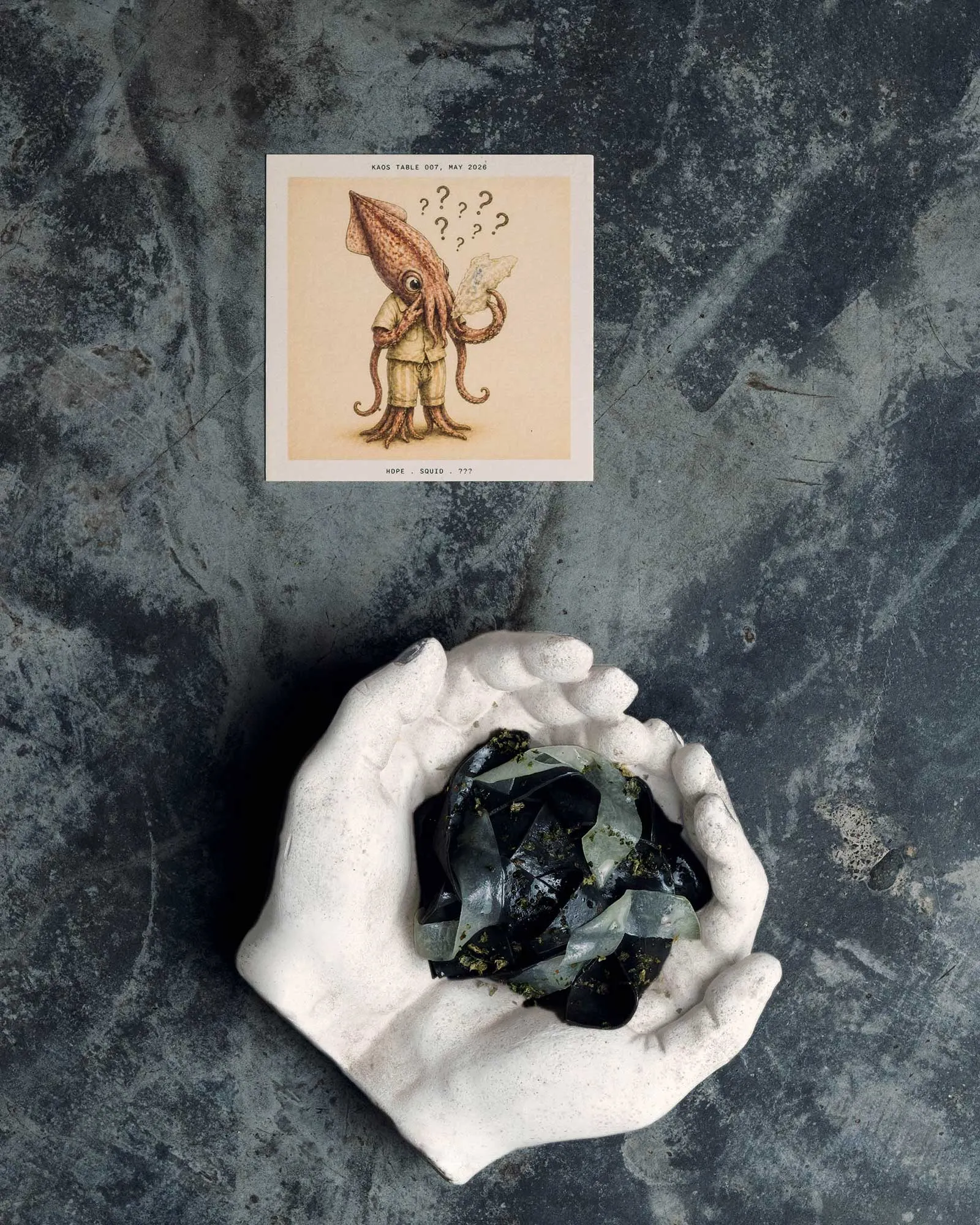White ceramic hand-shaped bowl filled with jagged dark-green glass shards sits on a textured dark surface, with a small illustrated card nearby.