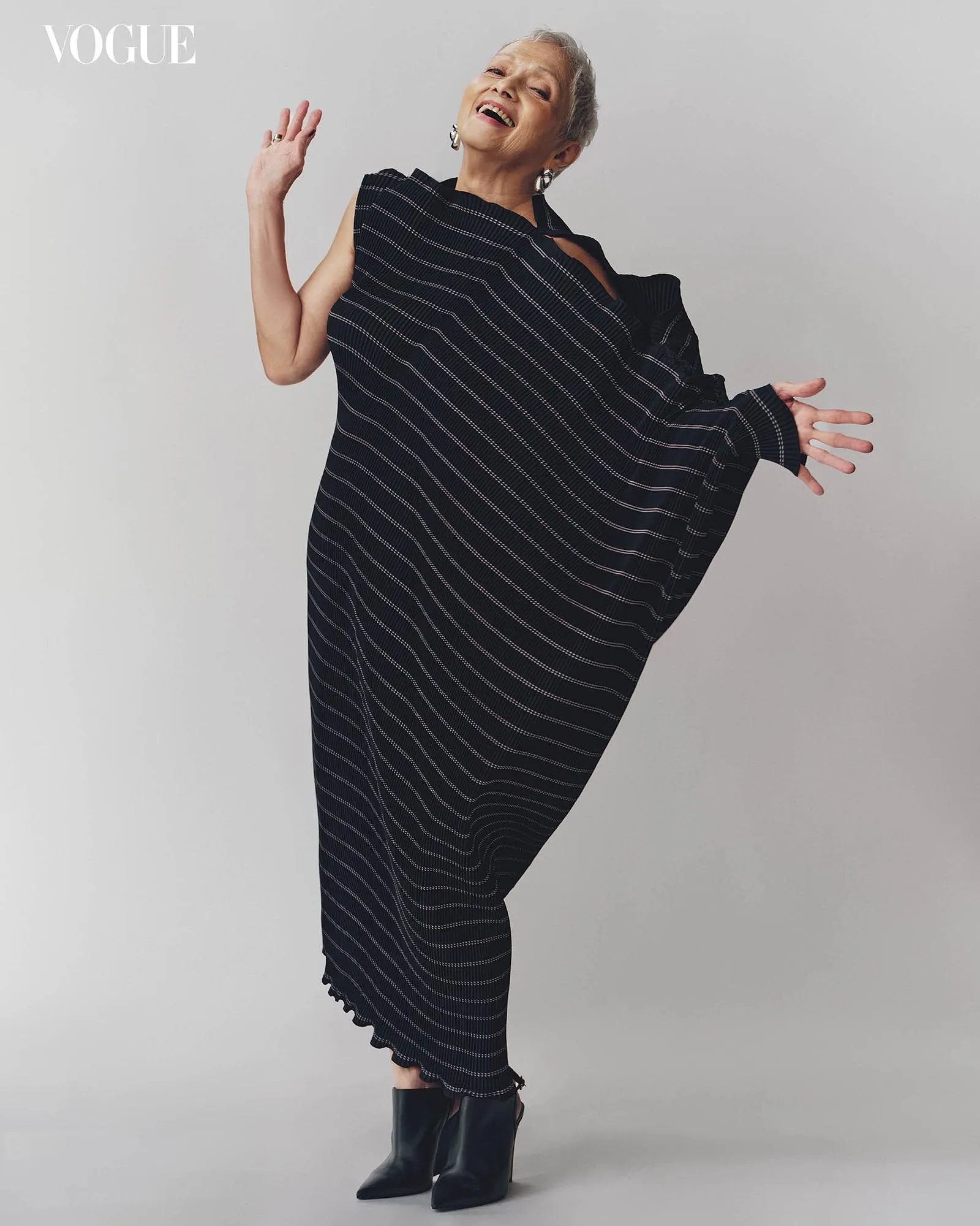Vogue logo in the corner; cheerful older woman wearing a black striped poncho and black boots, posing playfully against a gray backdrop.