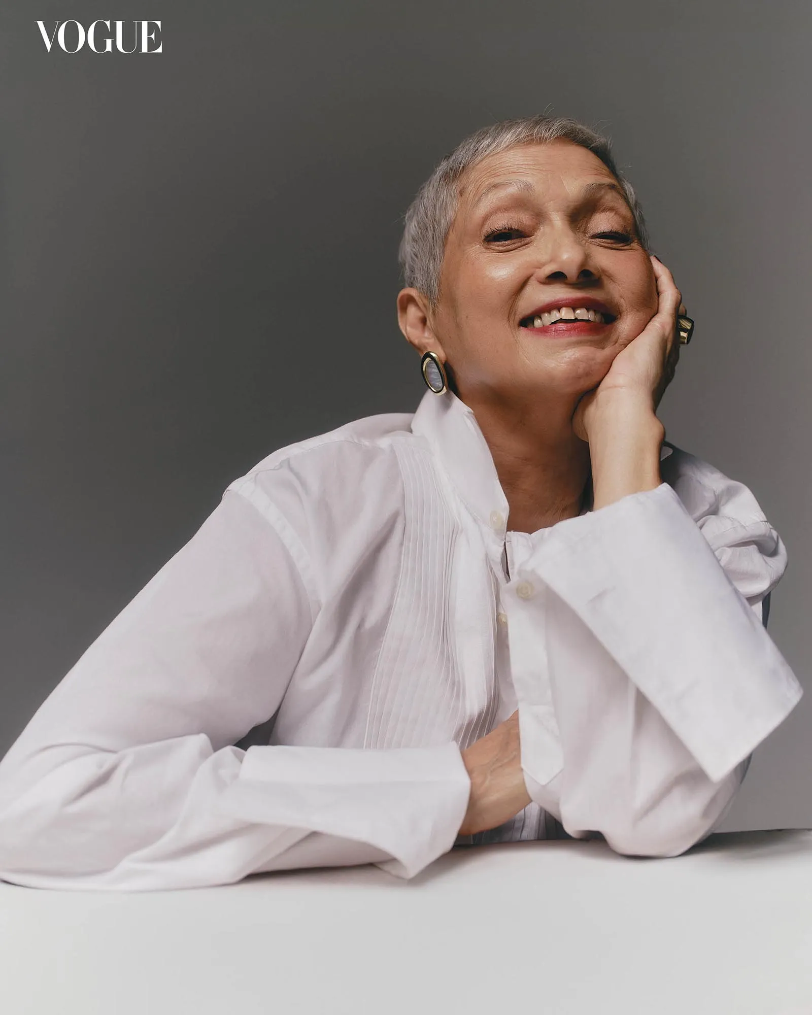 Smiling older woman with short gray hair in a white blouse, resting her cheek on her hand; Vogue logo in the top-left corner.