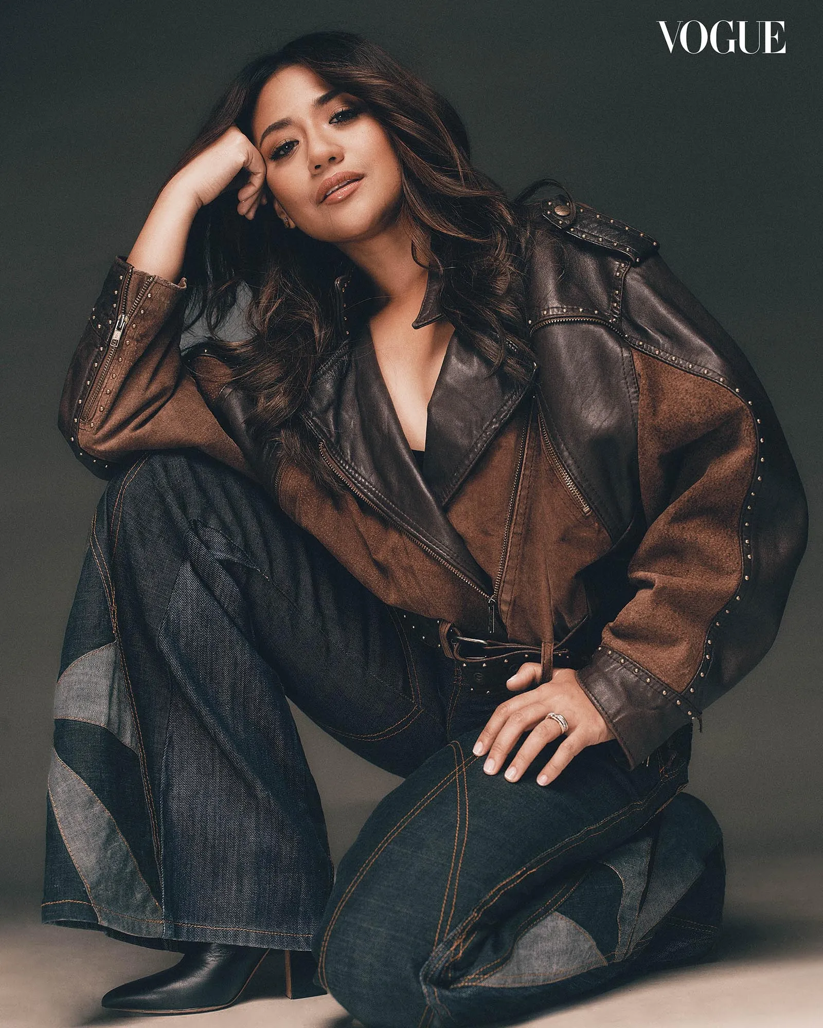 Editorial portrait of a female model in a brown leather jacket and dark denim, posed with hand to face; Vogue logo in the corner.