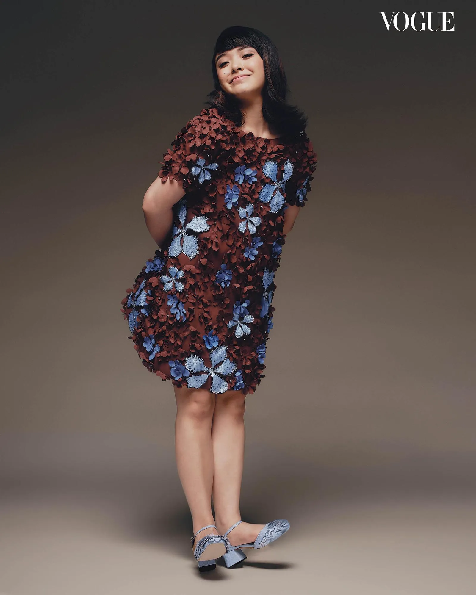 Model wearing a burgundy dress covered in blue flower appliqués, standing with a slight smile against a brown backdrop; Vogue logo in top-right corner.