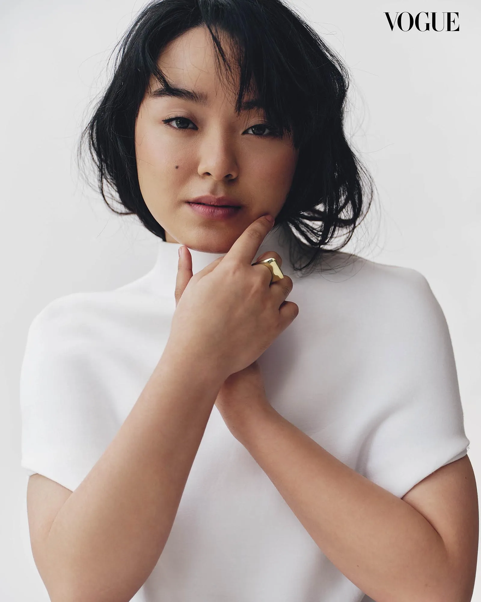 Close-up portrait of a woman with dark hair in a white top, hands gently touching her chin; Vogue logo in the top-right corner.