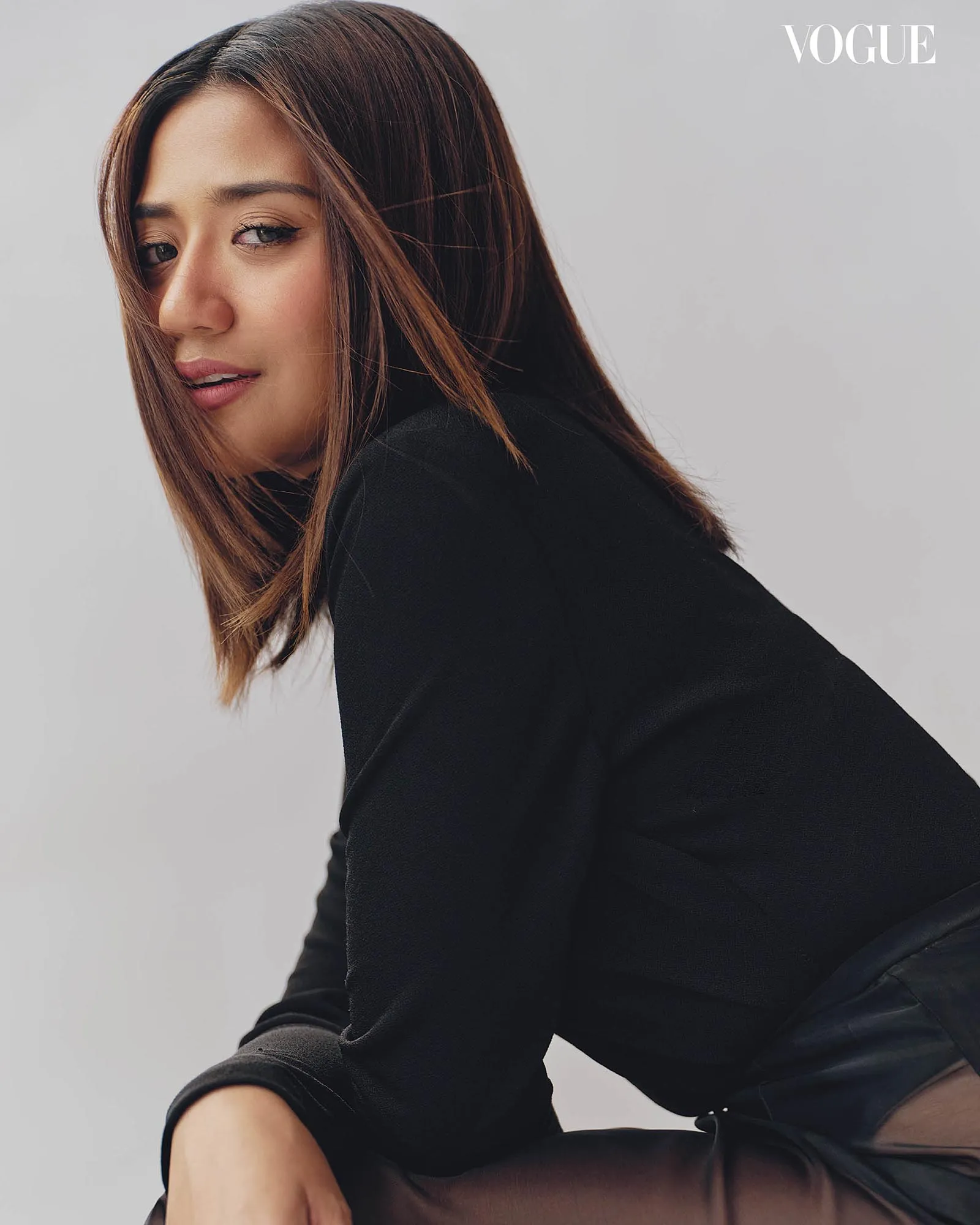 Close-up portrait of a young woman with shoulder-length light brown hair, wearing a black top, looking over her shoulder against a pale gray background; Vogue logo in the corner.