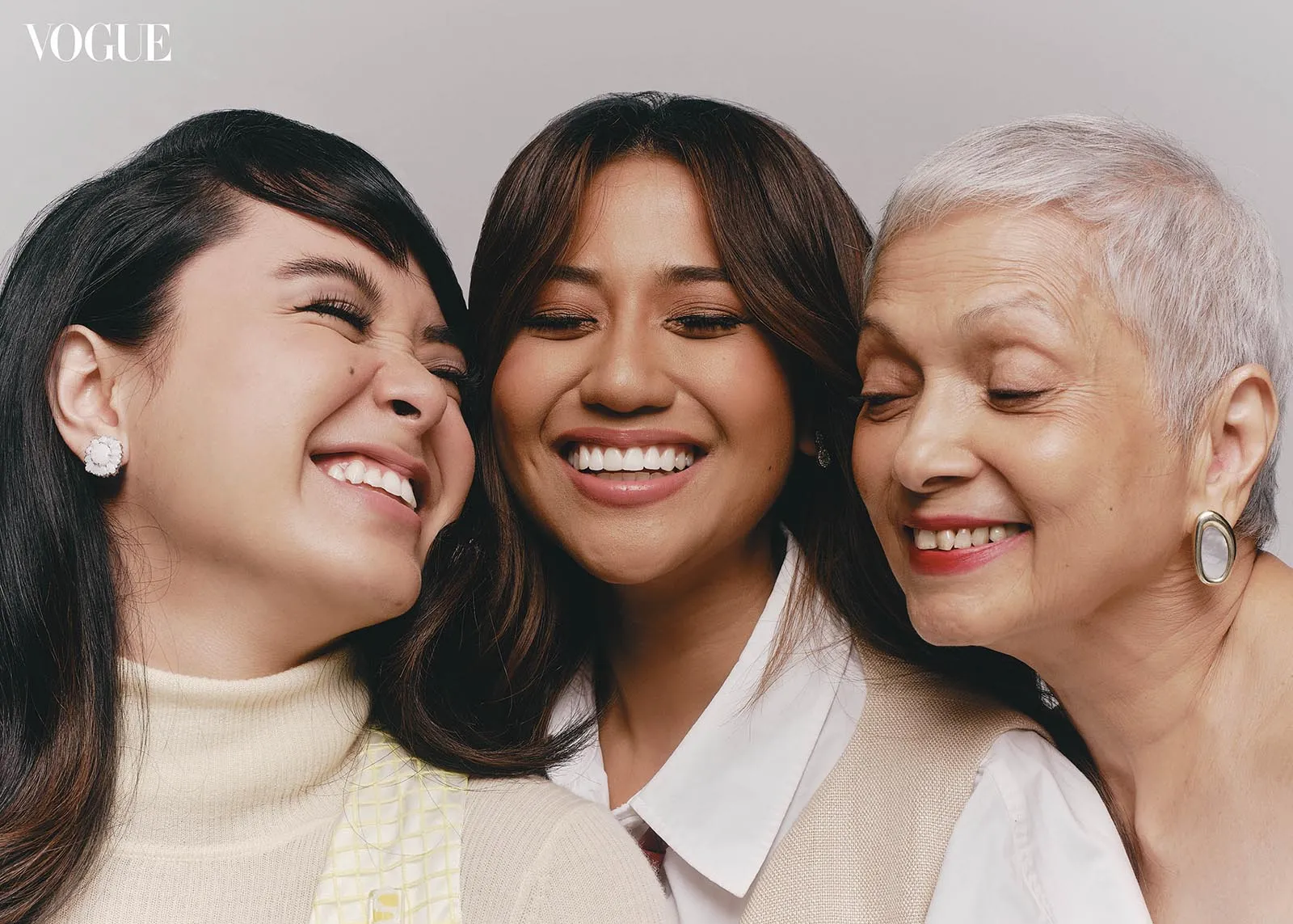 Three women of different ages smiling together, close‑up portrait, Vogue logo in the top-left corner.
