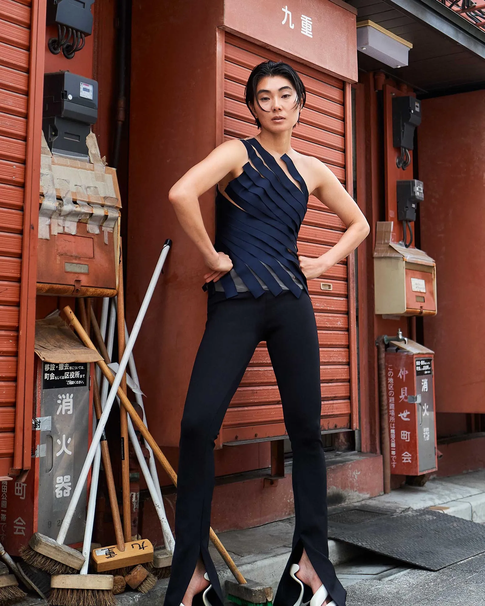 Woman in a navy, cutout pleated top and black pants poses with hands on hips outside a red storefront with Japanese signage and tools nearby.