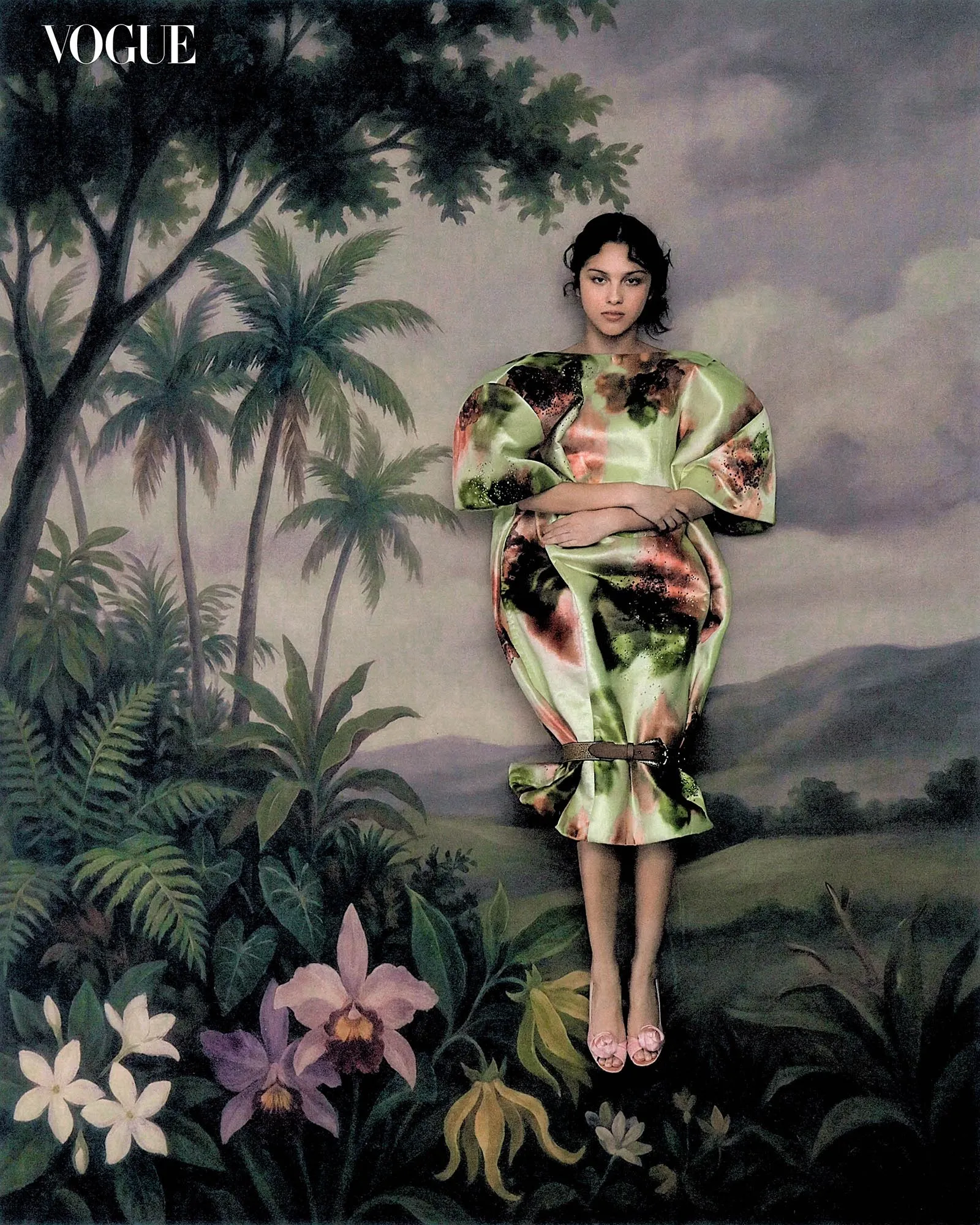 Fashion editorial image: a woman in a green floral dress stands among lush tropical plants on a painterly jungle backdrop, with a calm, poised expression.