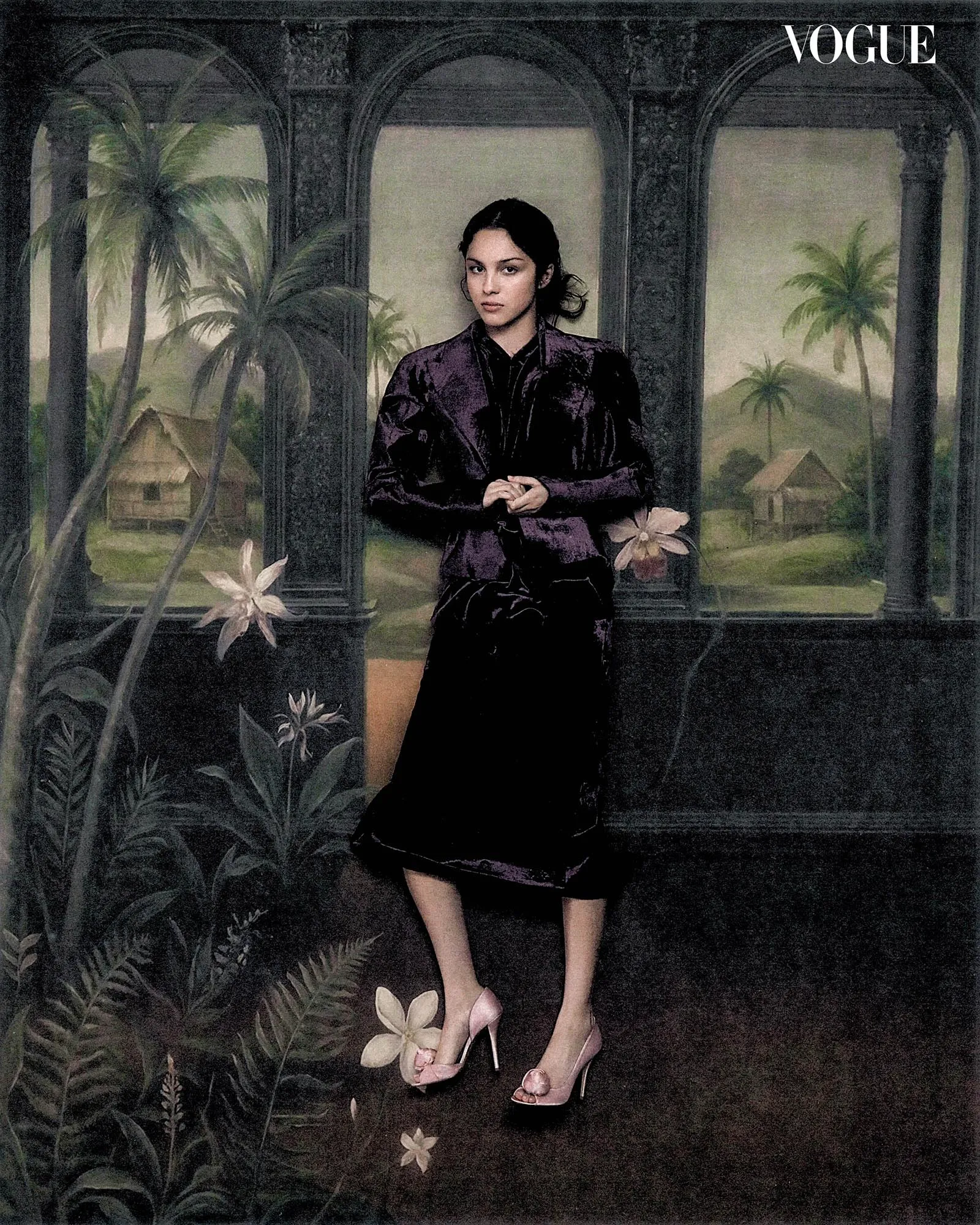 Fashion model in a dark purple velvet suit stands with hands clasped in front, posed against a tropical painted backdrop (Vogue watermark).