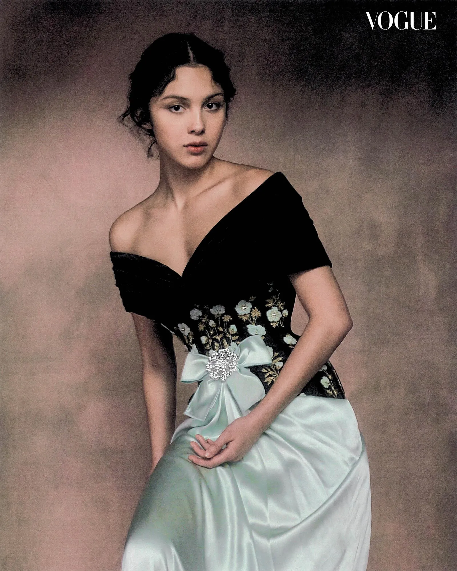 Woman in an off-the-shoulder black gown with floral embroidery and a pale blue sash-like skirt, posing against a neutral backdrop, Vogue logo visible in the corner.