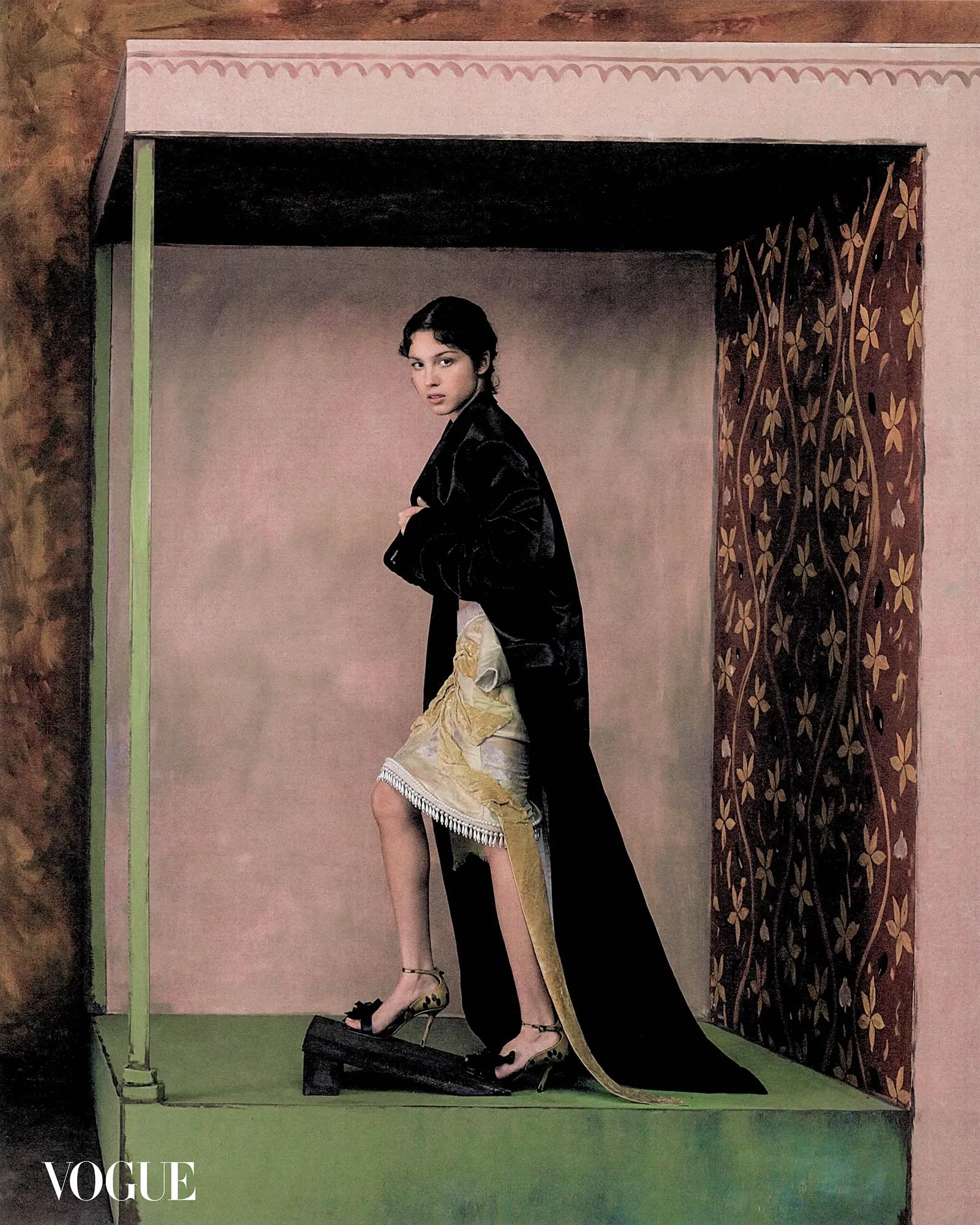 Vogue magazine cover featuring a model in a black cape and gold dress standing on a green platform inside a pink, decorative studio set with a floral-patterned curtain to the right.