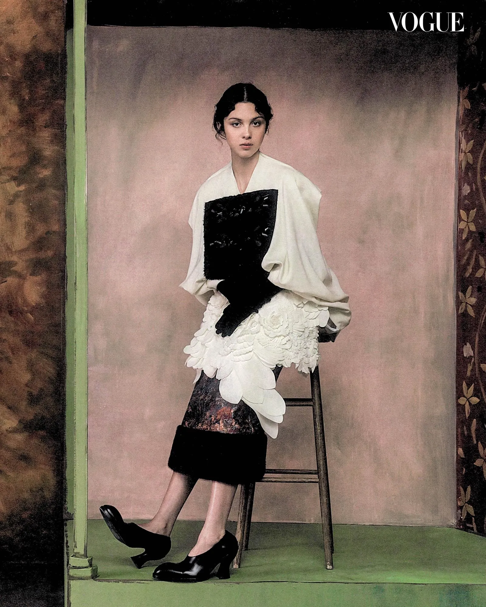 Editorial fashion shot: model in a white, sculptural dress with black panel, standing on a chair against a muted backdrop (Vogue logo in corner).