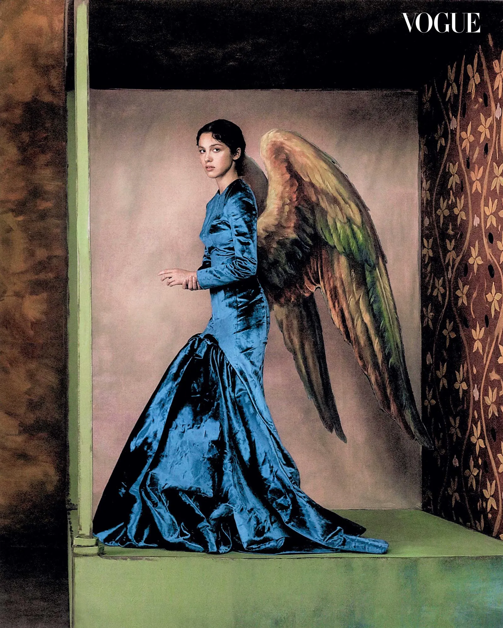 Woman in a blue satin gown stands in a doorway with large multicolored wings extending from her back, in a vintage-room setting.