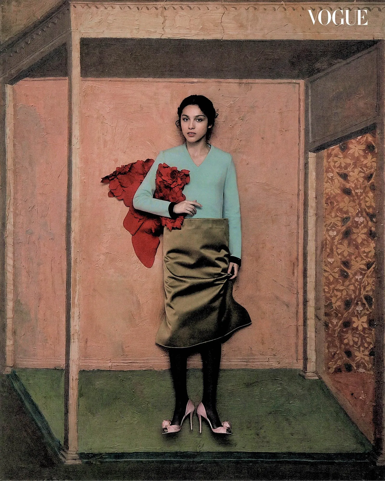 Fashion editorial cover: woman in a pastel mint top and satin olive skirt, holding a red bouquet, standing in a pink-walled room.