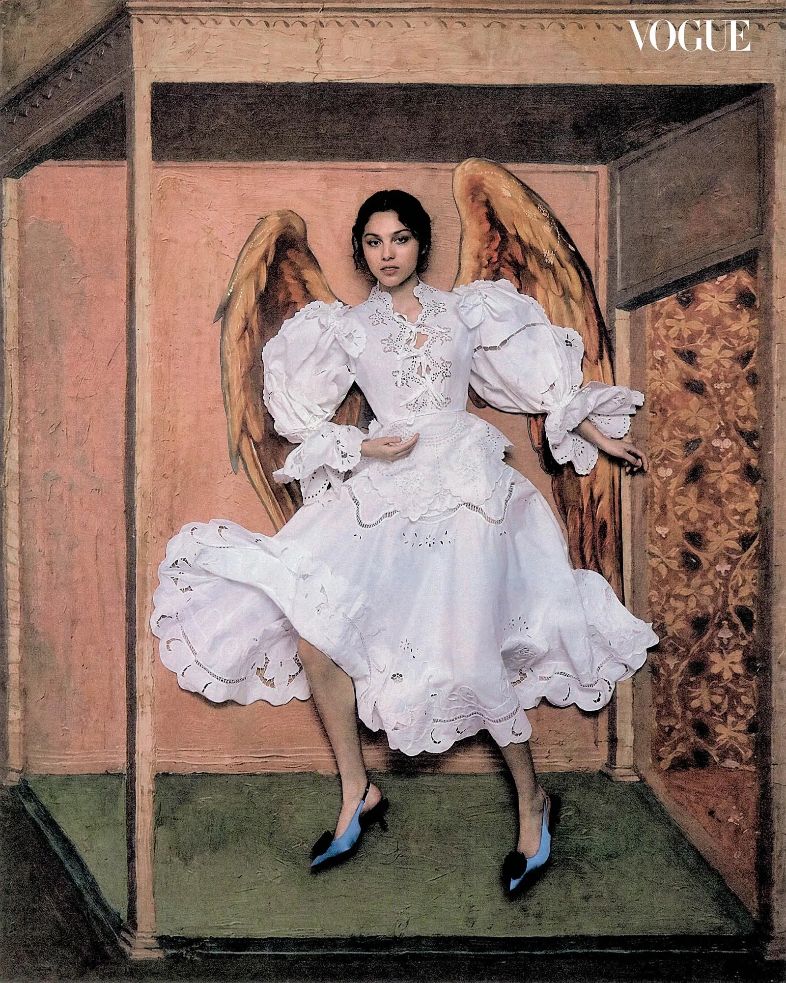 Portrait of a woman in a white lace gown with large angel wings, standing in a pink-toned stage frame; Vogue logo in the top-right corner.