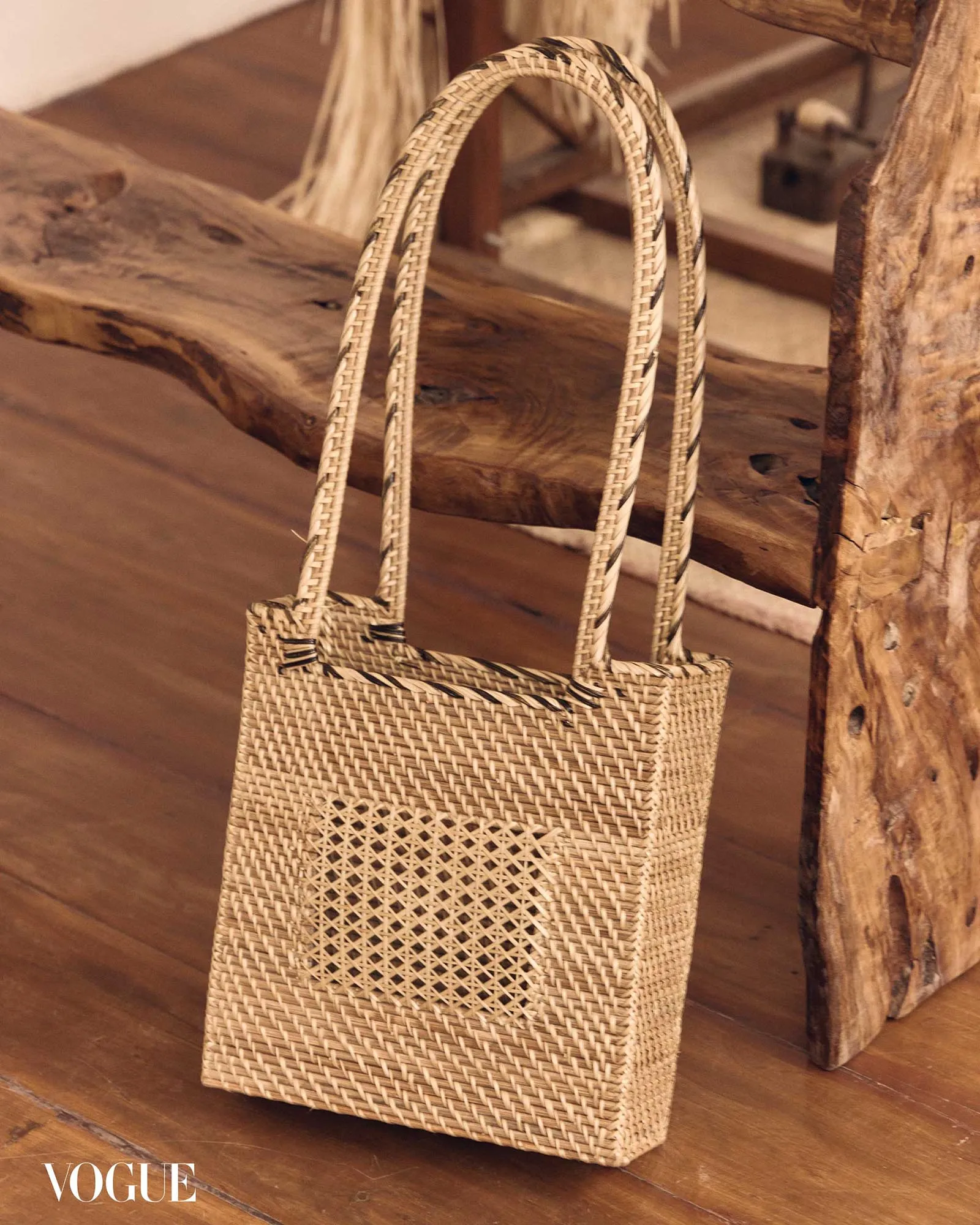Beige woven straw handbag with two handles resting on a wooden floor, Vogue watermark visible in the corner.