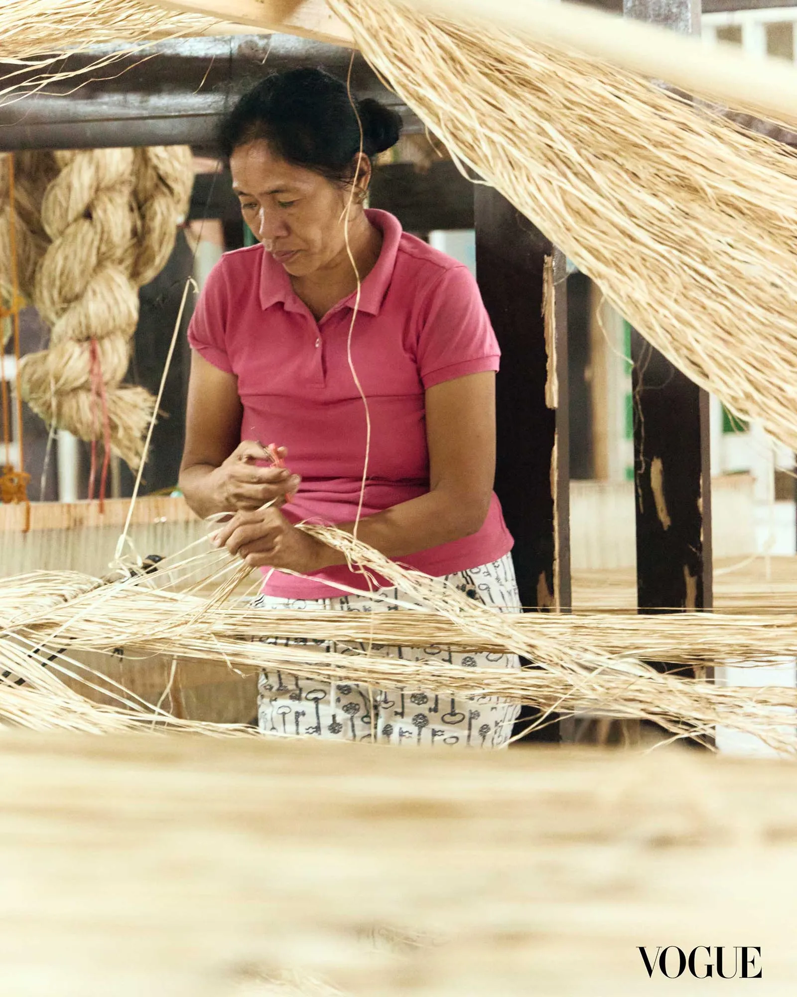 Woman in a pink polo weaving straw fibers in a workshop, with large bundles of straw surrounding her; Vogue logo appears in the bottom right.