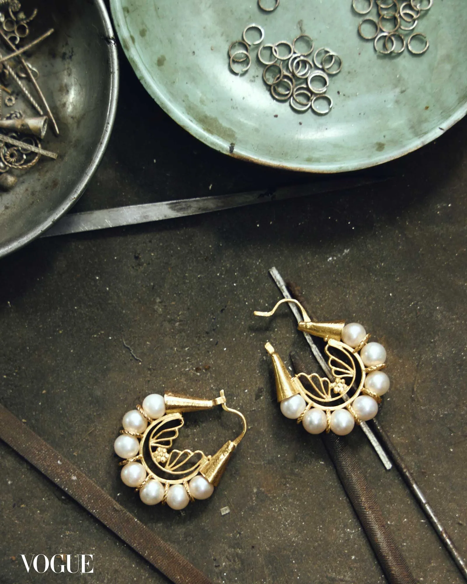 Gold-tone jewelry with white pearls arranged on a dark surface, Vogue watermark visible in the corner.