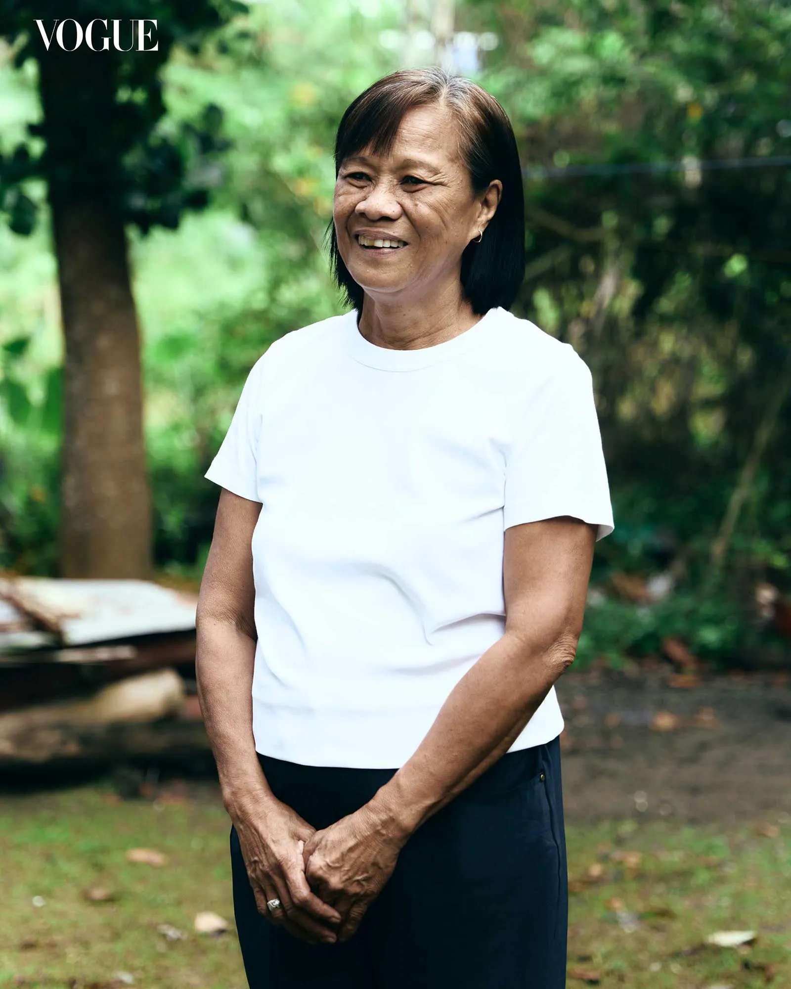 Older woman standing outdoors in a white shirt, smiling, hands clasped in front, greenery in background (Vogue in corner).