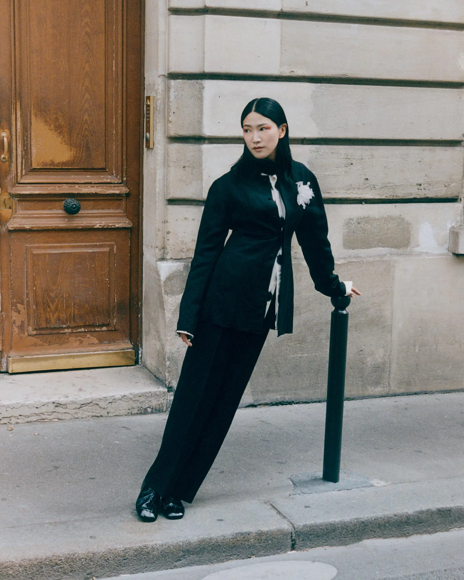 Woman in a black suit stands on a city sidewalk, leaning against a green post beside a stone wall and wooden door.