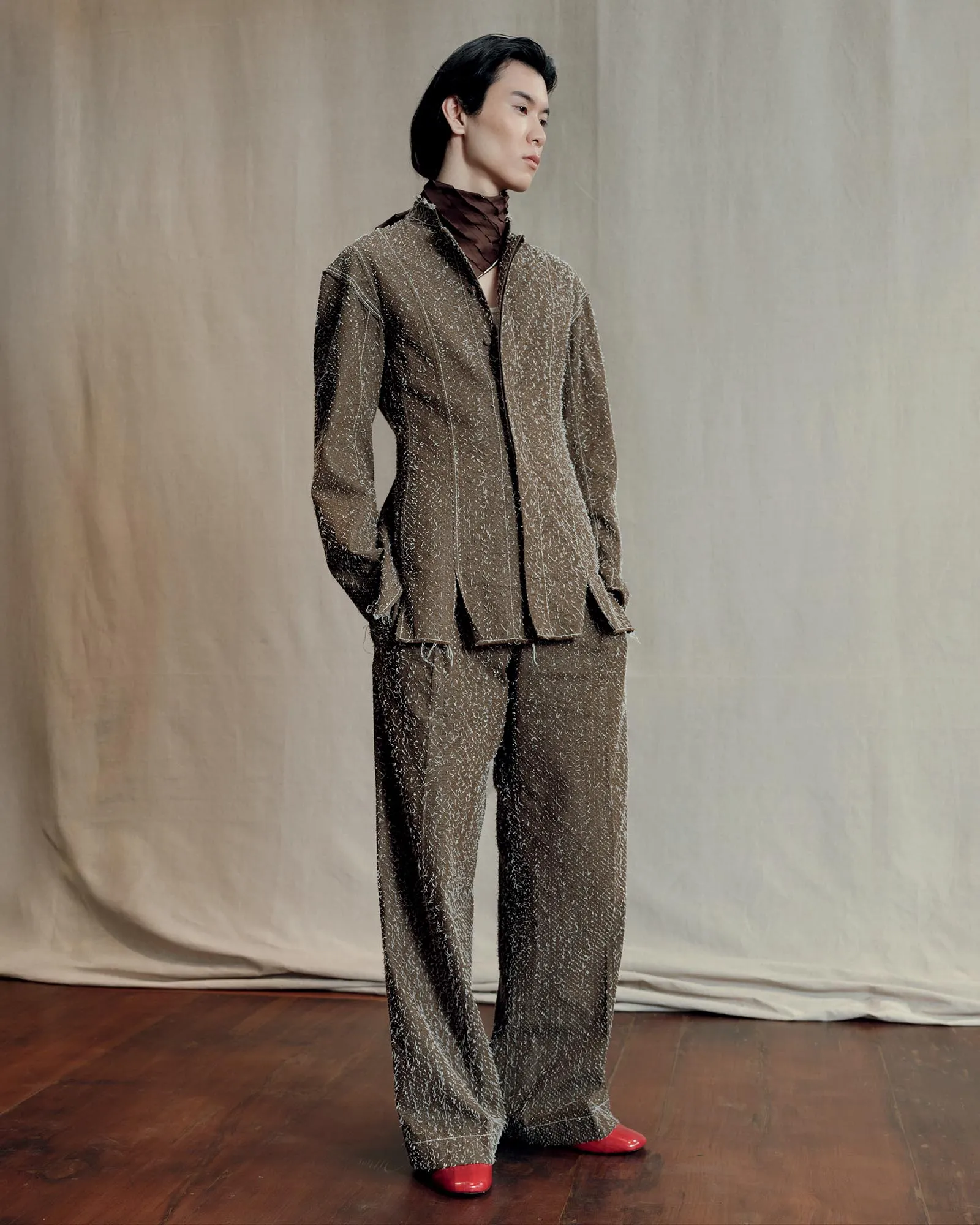 Model wearing a speckled brown suit with wide-legged pants, standing on a wooden floor against a neutral backdrop, red shoes visible at the hem.