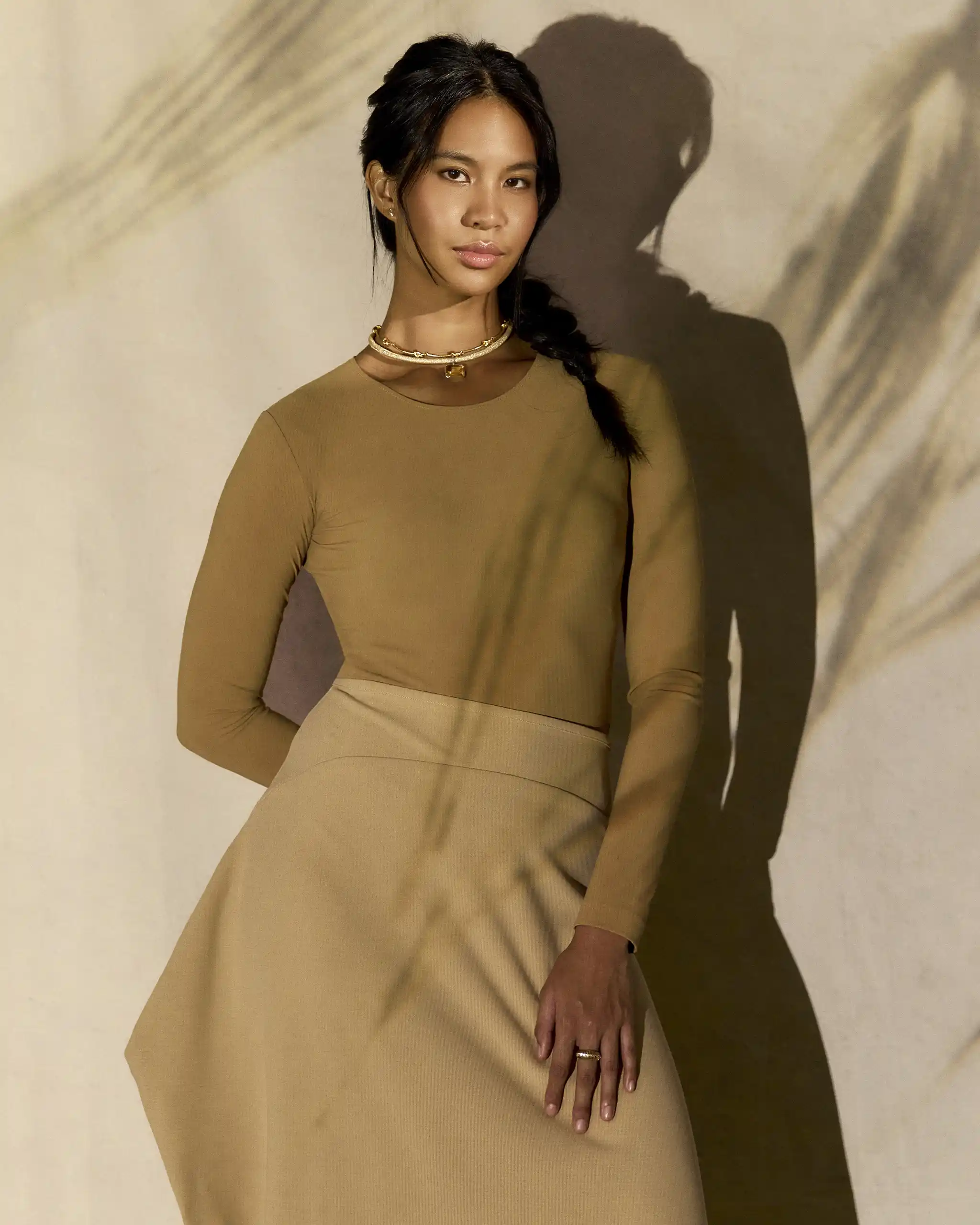 Woman in a beige long-sleeve dress with a gold necklace, standing against a shadowy beige backdrop.