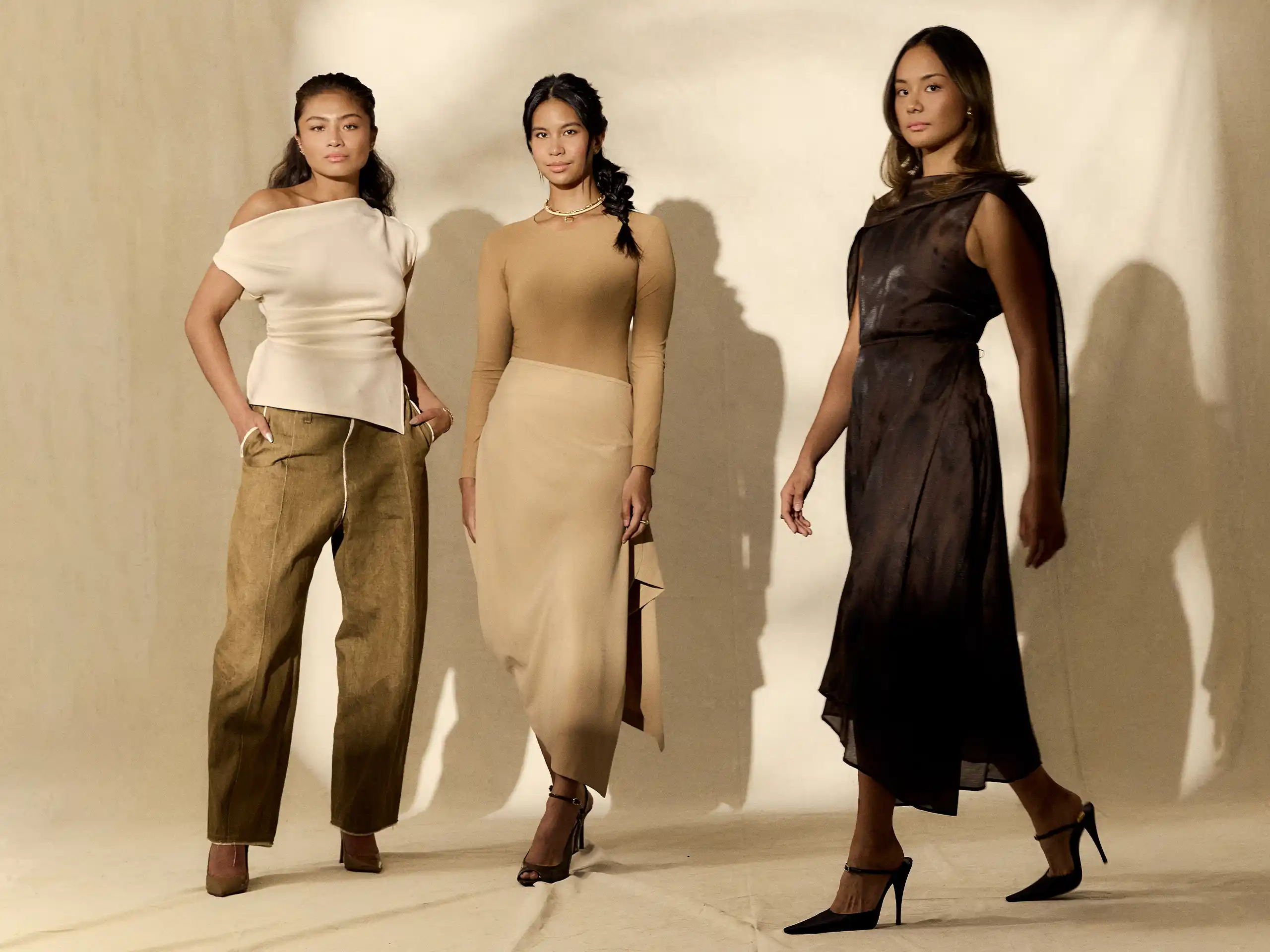 Three female models pose in a fashion studio wearing neutral-toned outfits: white top with tan pants, beige dress, and a dark sheer gown.