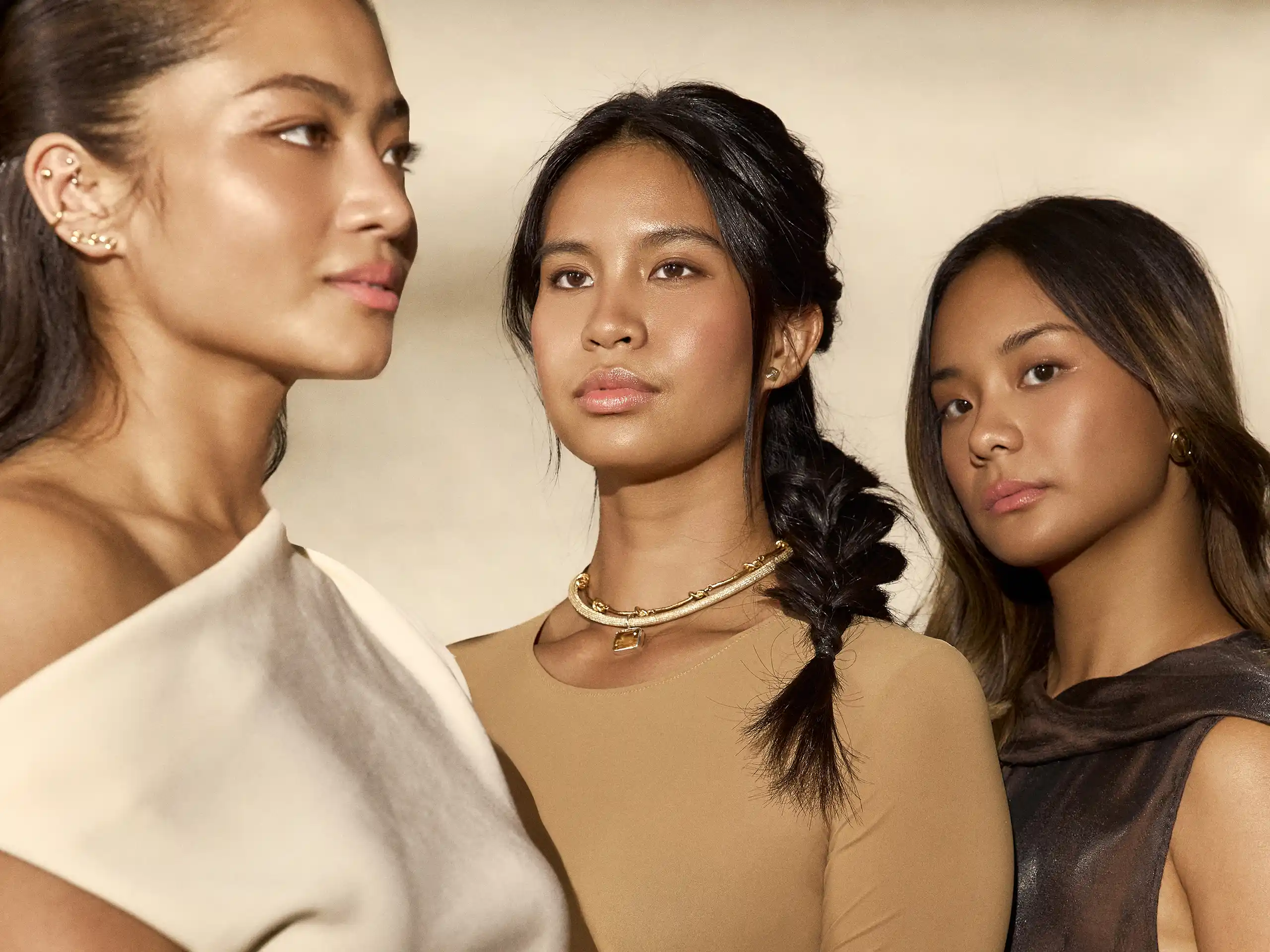 Three women posing closely together; center wears a beige top with gold necklace, others in white and dark outfits with jewelry.