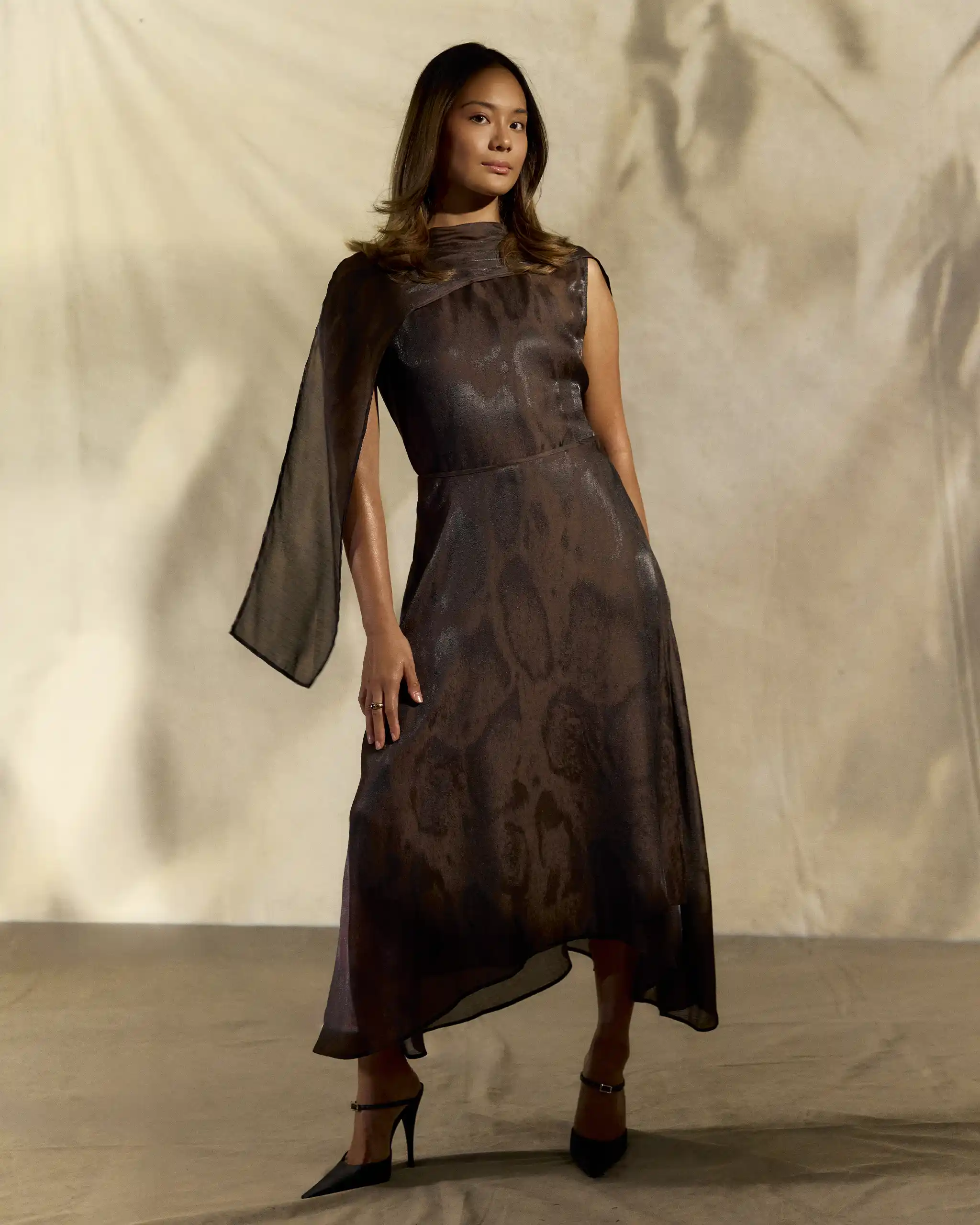 Woman in a dark metallic, one-shoulder gown standing on a beige backdrop, mid-step with heels visible.