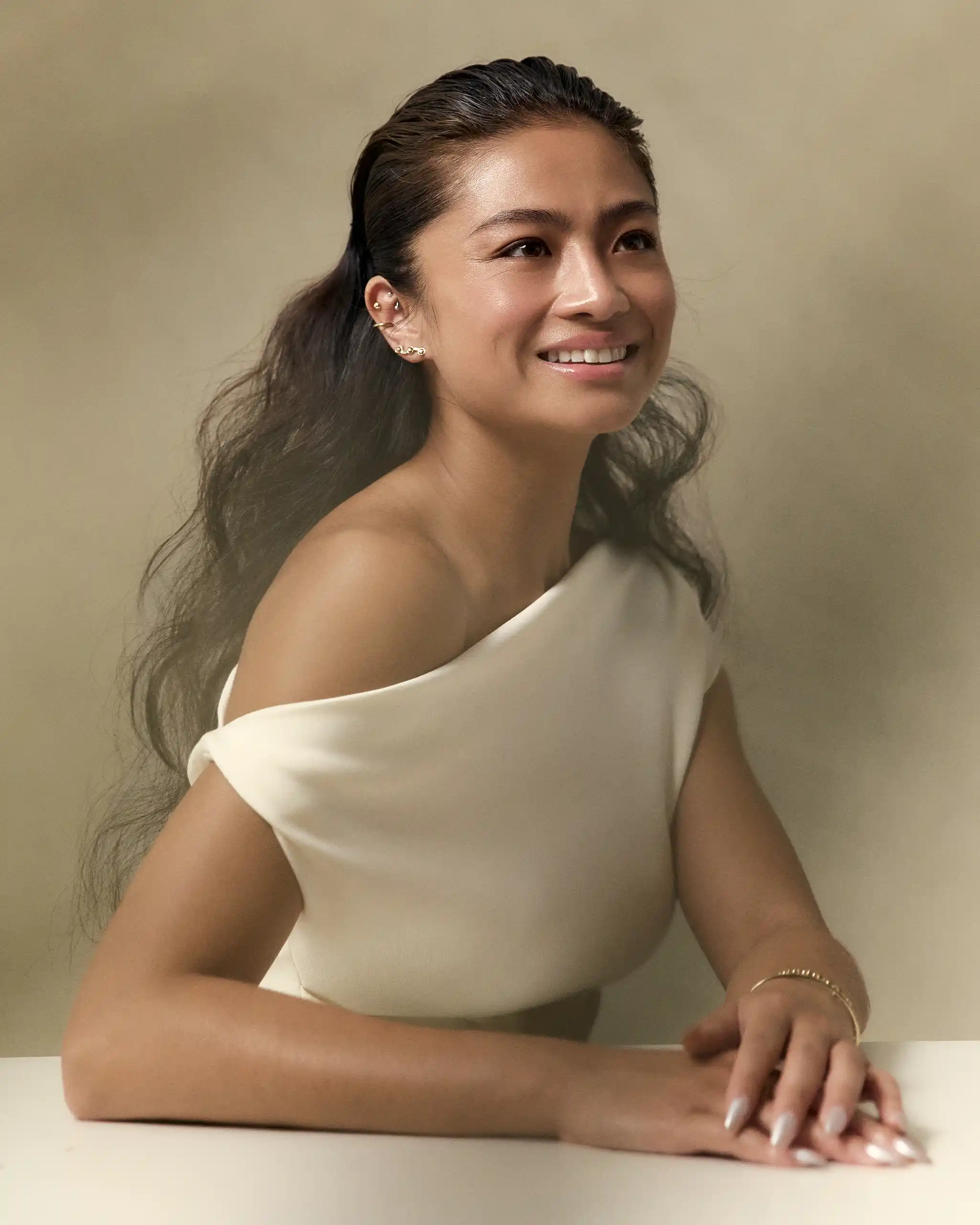 Smiling woman with dark hair tied back rests her arms on a white table, wearing a white off-shoulder top and gold jewelry against a beige background.