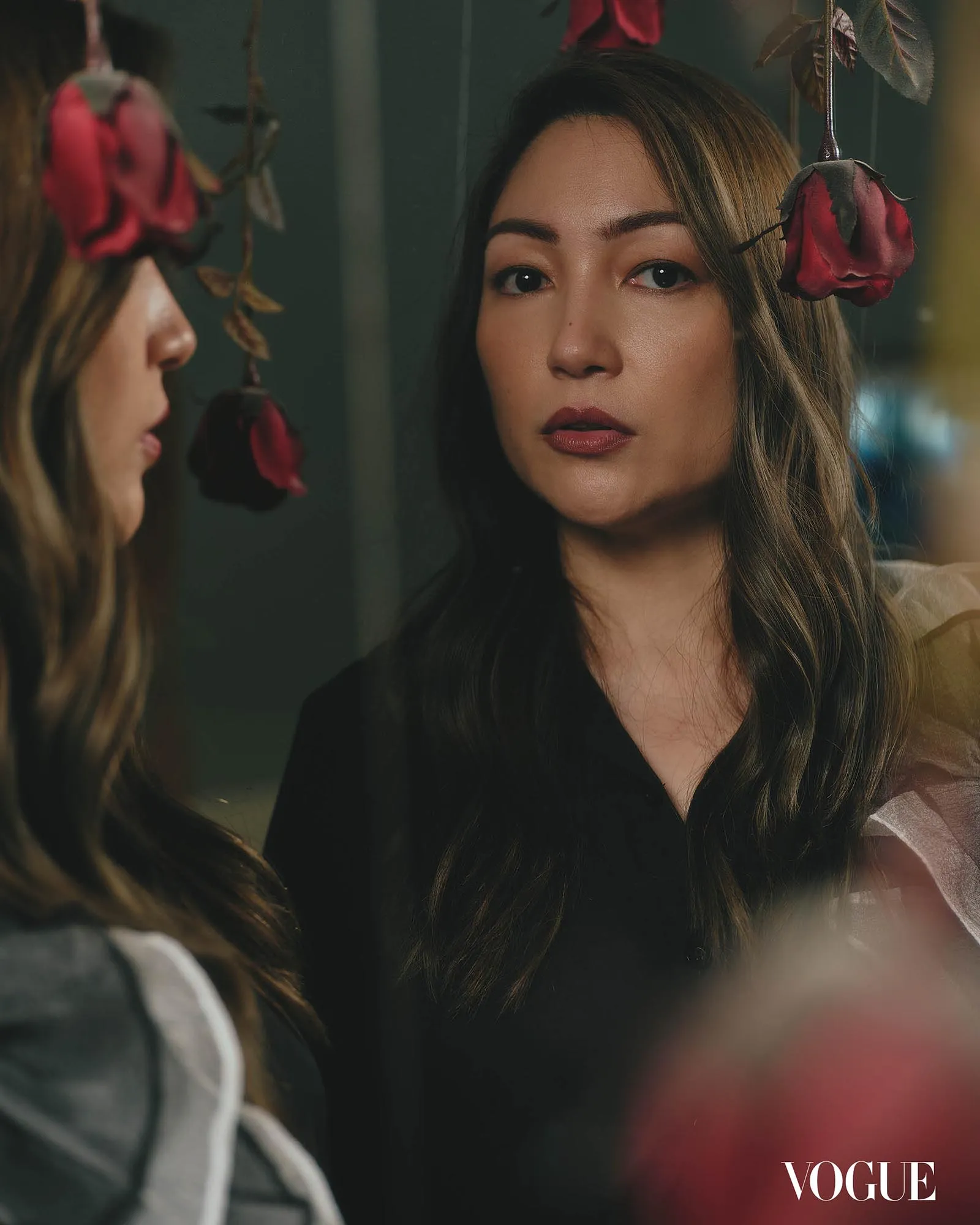 Close-up of a woman looking into a mirror with red roses hanging nearby; Vogue logo visible in the corner.