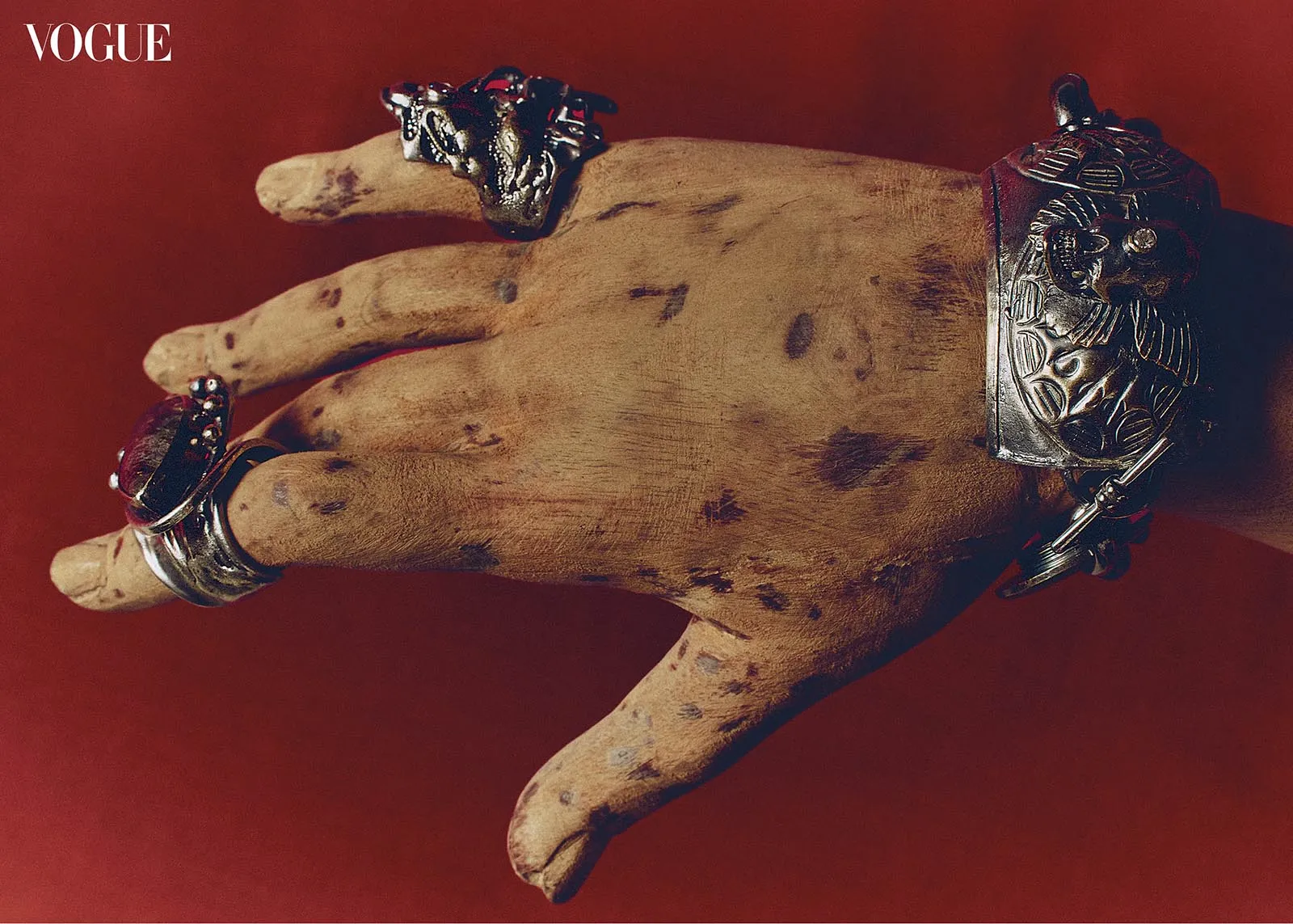 Decorative hand wearing multiple silver rings and a large ornate cuff against a red background; Vogue logo visible in the corner.