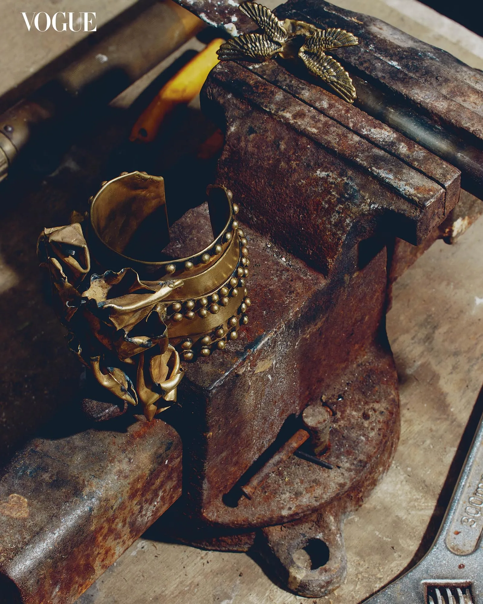 Gold bracelets and a beaded necklace draped over a rusted anvil with Vogue logo in the corner.