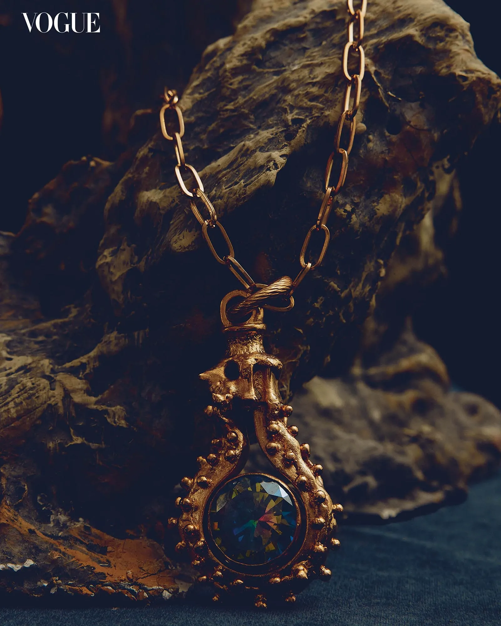 Close-up of a gold chain necklace with an ornate pendant and a faceted multicolored gem, draped against a dark rock surface with the Vogue logo in the corner.