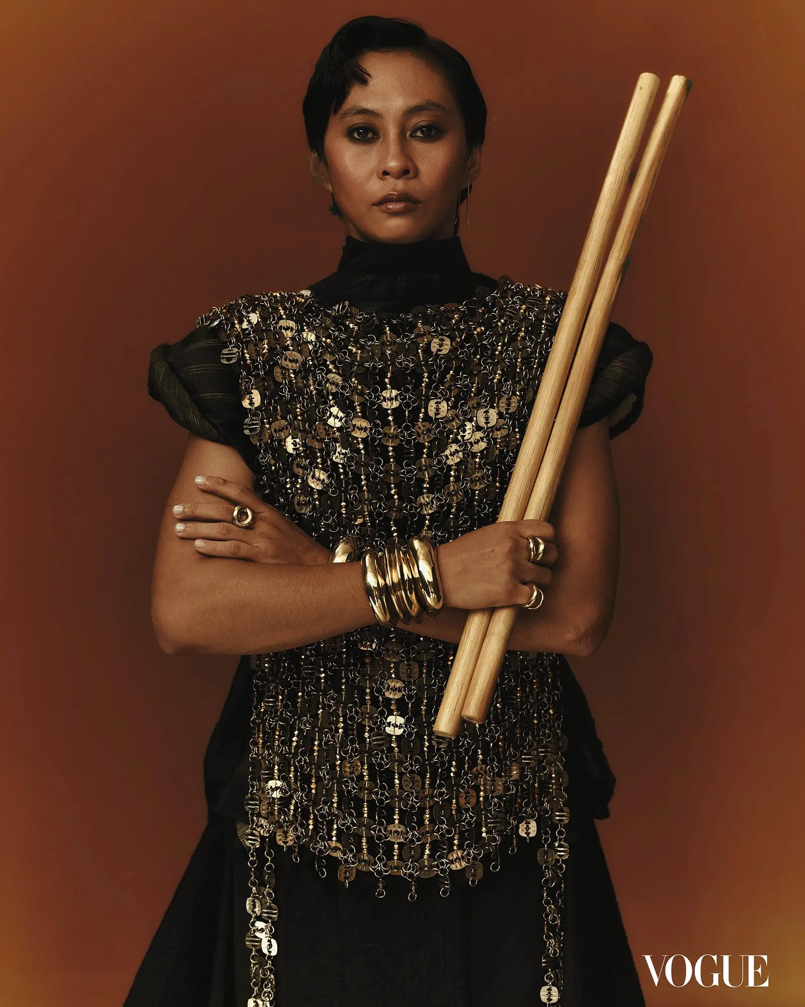 Portrait of a woman in a metallic, chainmail-style top holding two wooden drumsticks against a warm brown background; Vogue logo visible in the corner.