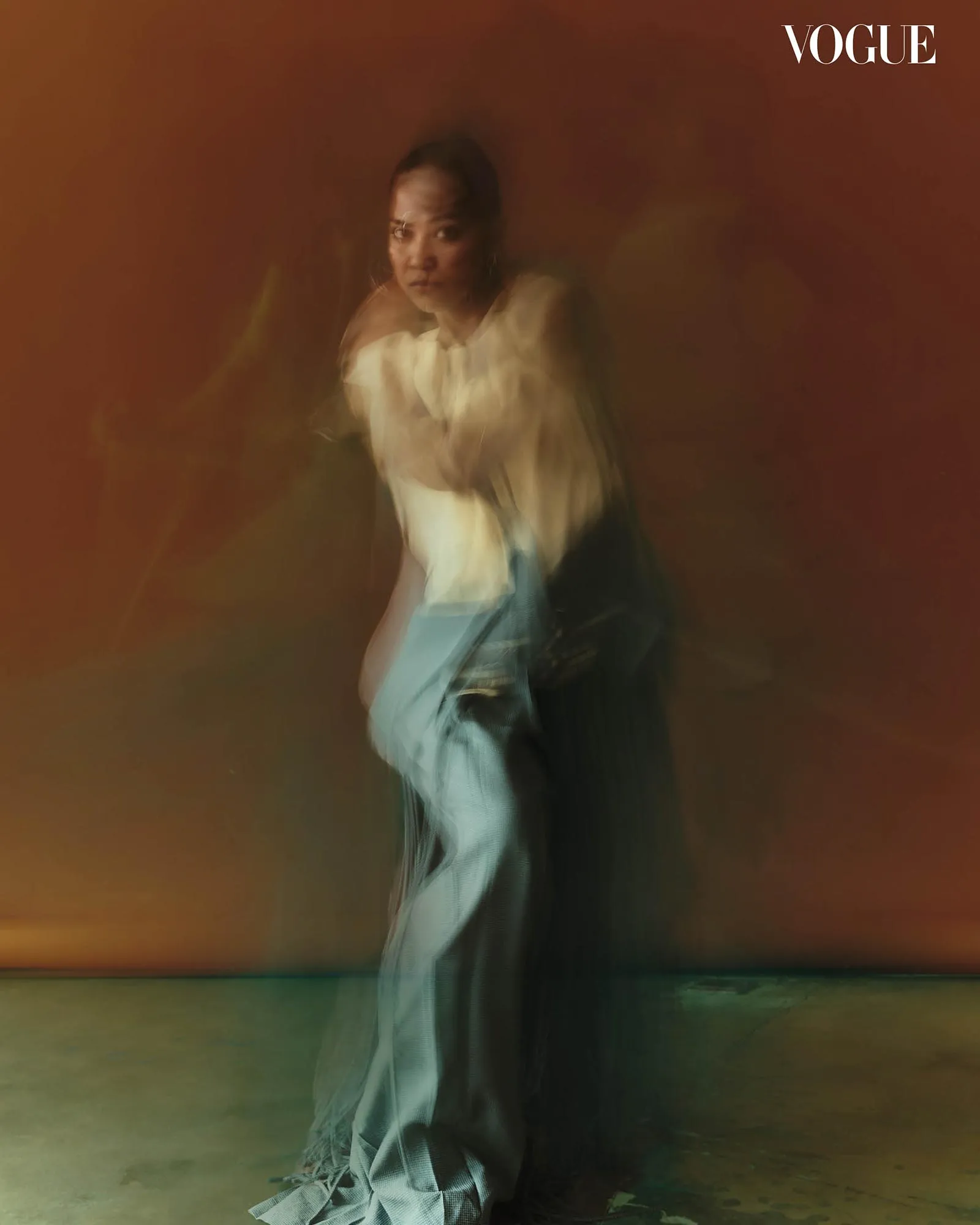 Blurry fashion portrait of a woman in a white blouse and jeans against an orange-green background, Vogue logo in the top-right corner.