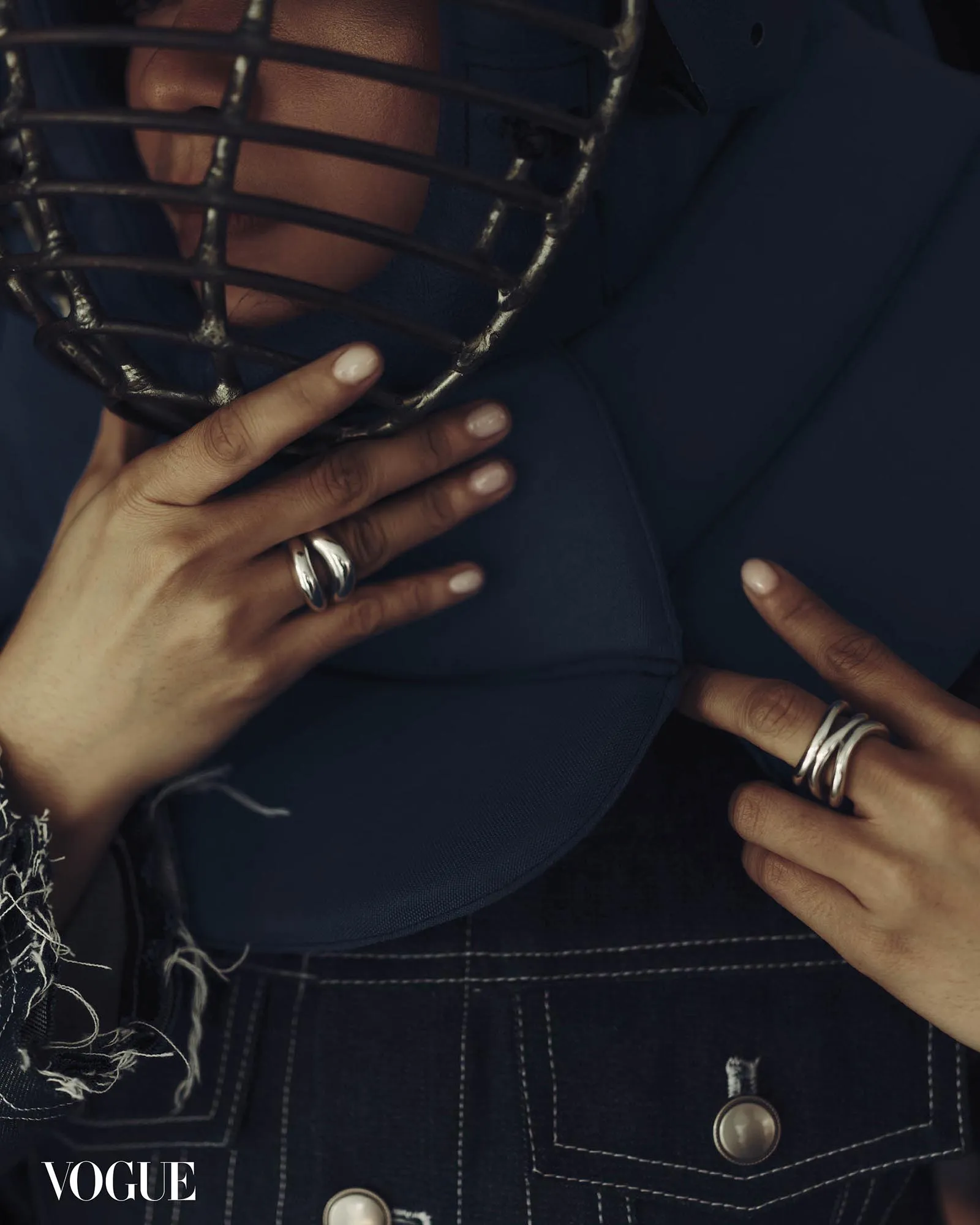 Close-up of hands holding a black metal helmet against a dark blue coat; Vogue logo visible.