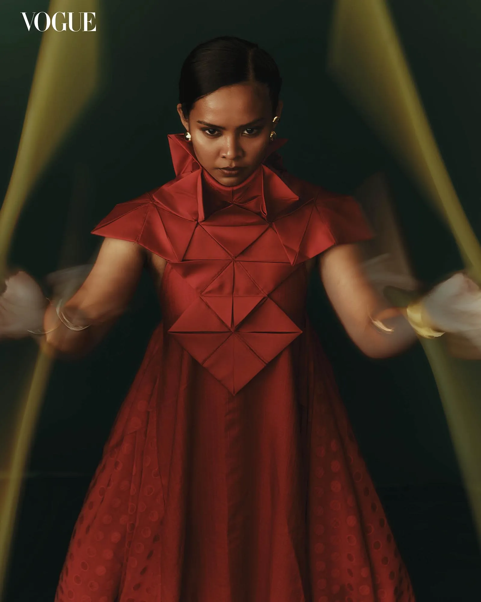 Editorial fashion portrait of a woman in a structured red dress, staring intently forward, Vogue logo top left.